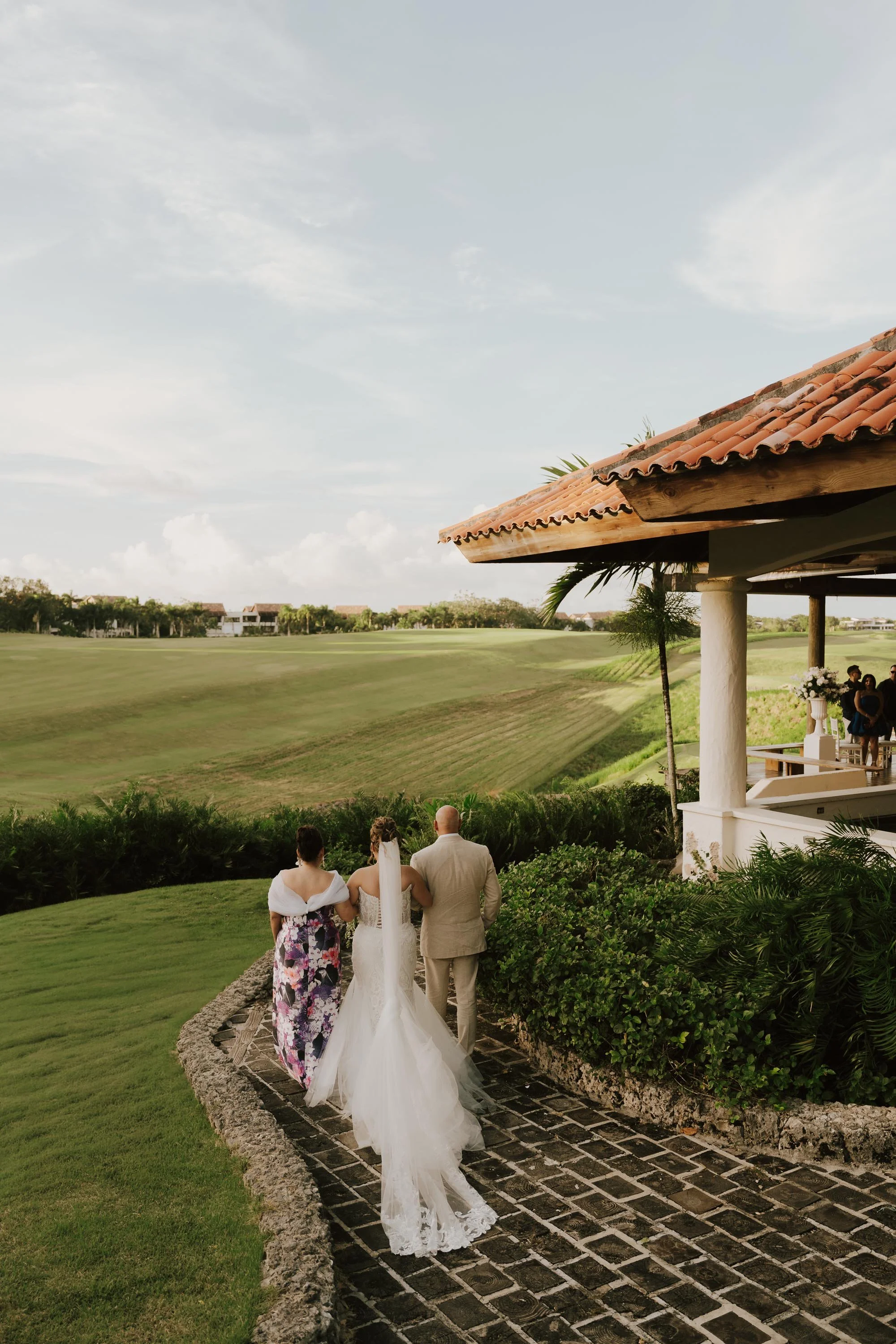 Bride escorted by her parents walking down the aisle during an outdoor wedding ceremony at Dye Fore, with a scenic golf course and landscape background.