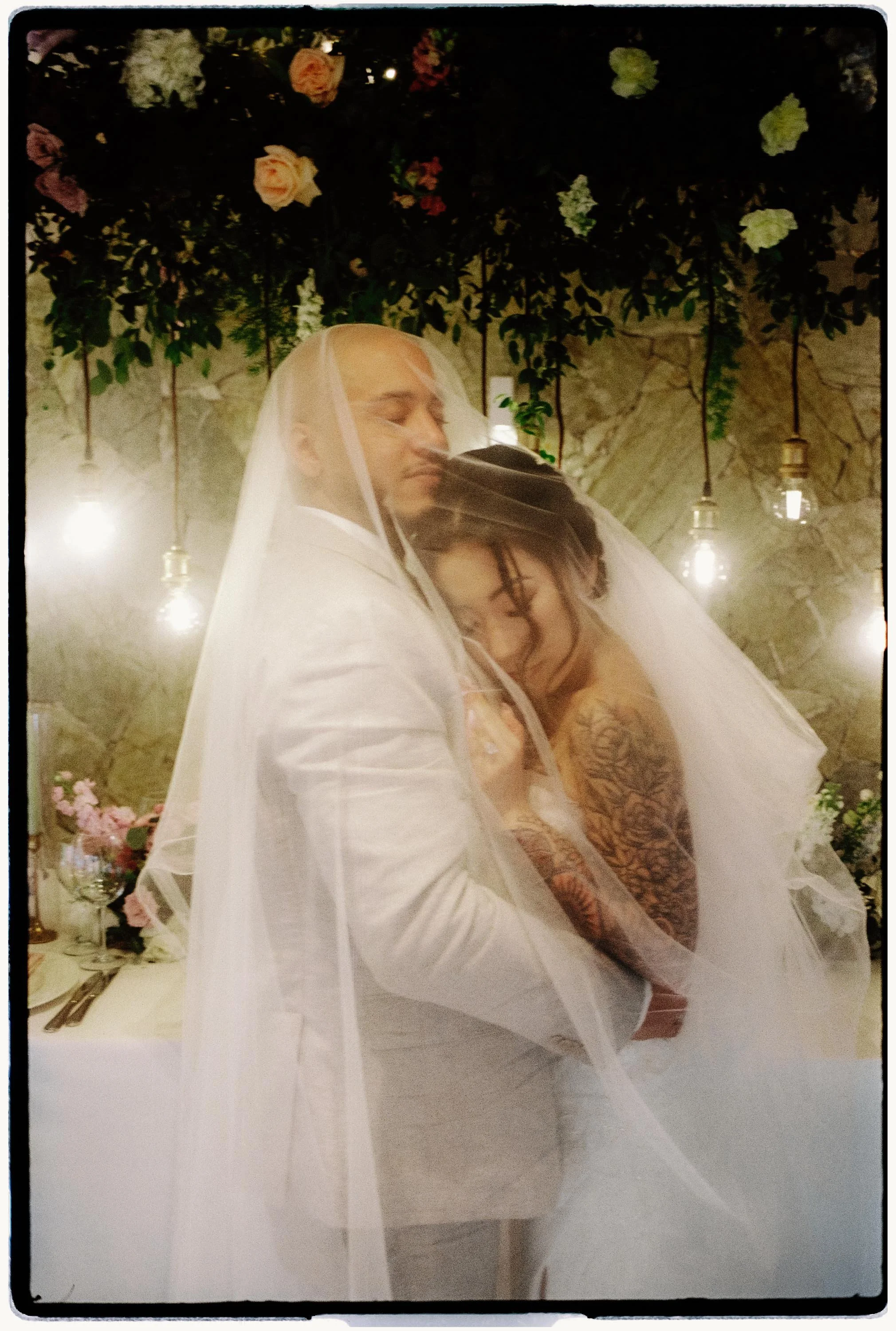 35mm film photo of a bride and groom embracing intimately under her wedding veil at their reception, illuminated by warm hanging Edison bulbs against a stone wall.