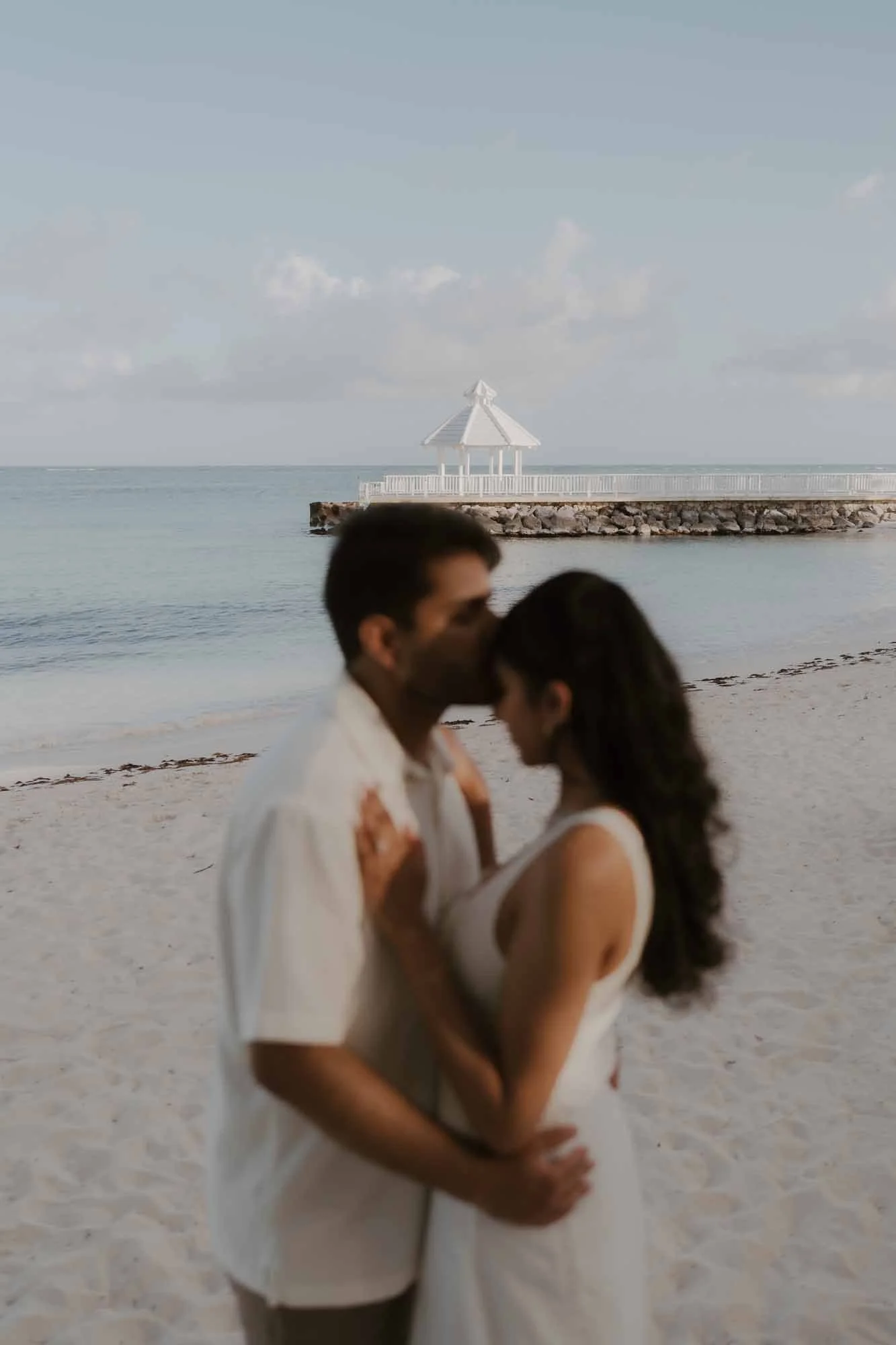Groom kissing the bride in the forehead while they are facing each other. In the background you can see a white gazebo and the clouds