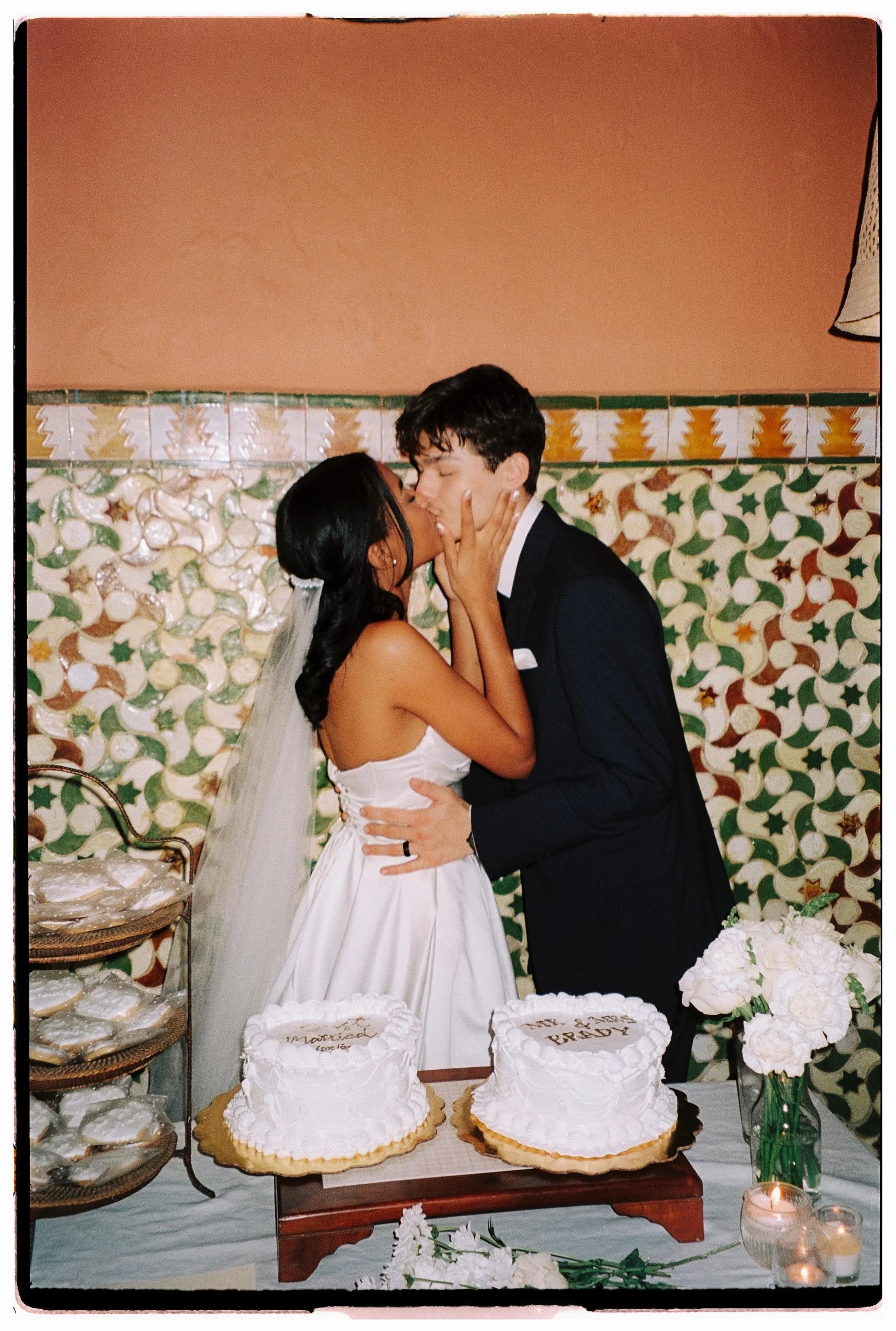 Bride and groom kissing behind two wedding cakes in front of a vintage tile wall at Microteatro.