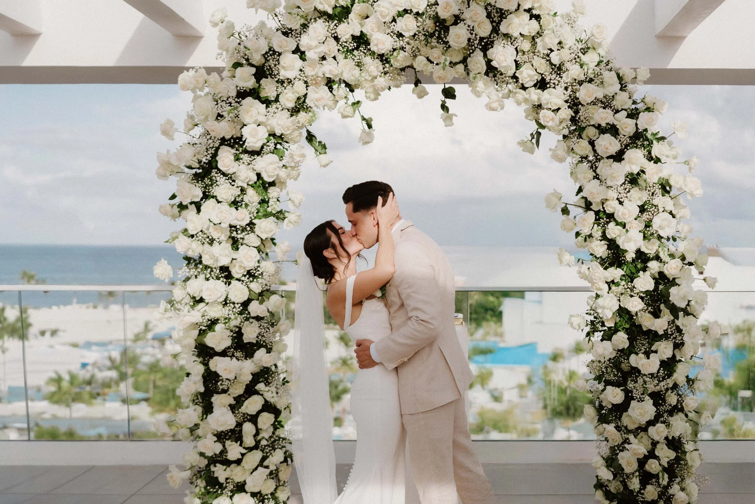 Bride and groom sharing a romantic first kiss under a lush white rose arch during their oceanfront destination wedding ceremony in Cap Cana Punta Cana