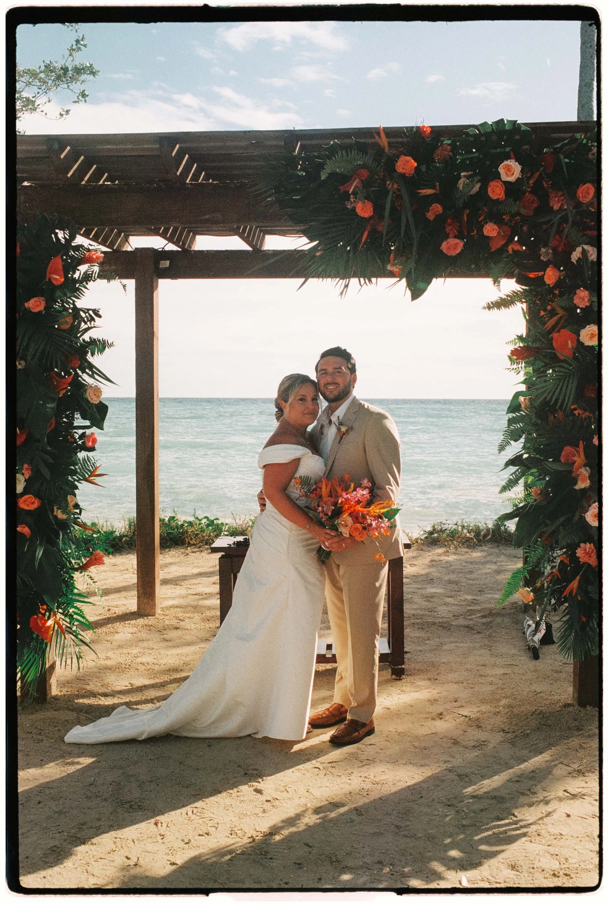 35 film mm photo of the couple standing and looking into the camera. The couple stands under a tropical flower arch and the beach is in the beackground.
