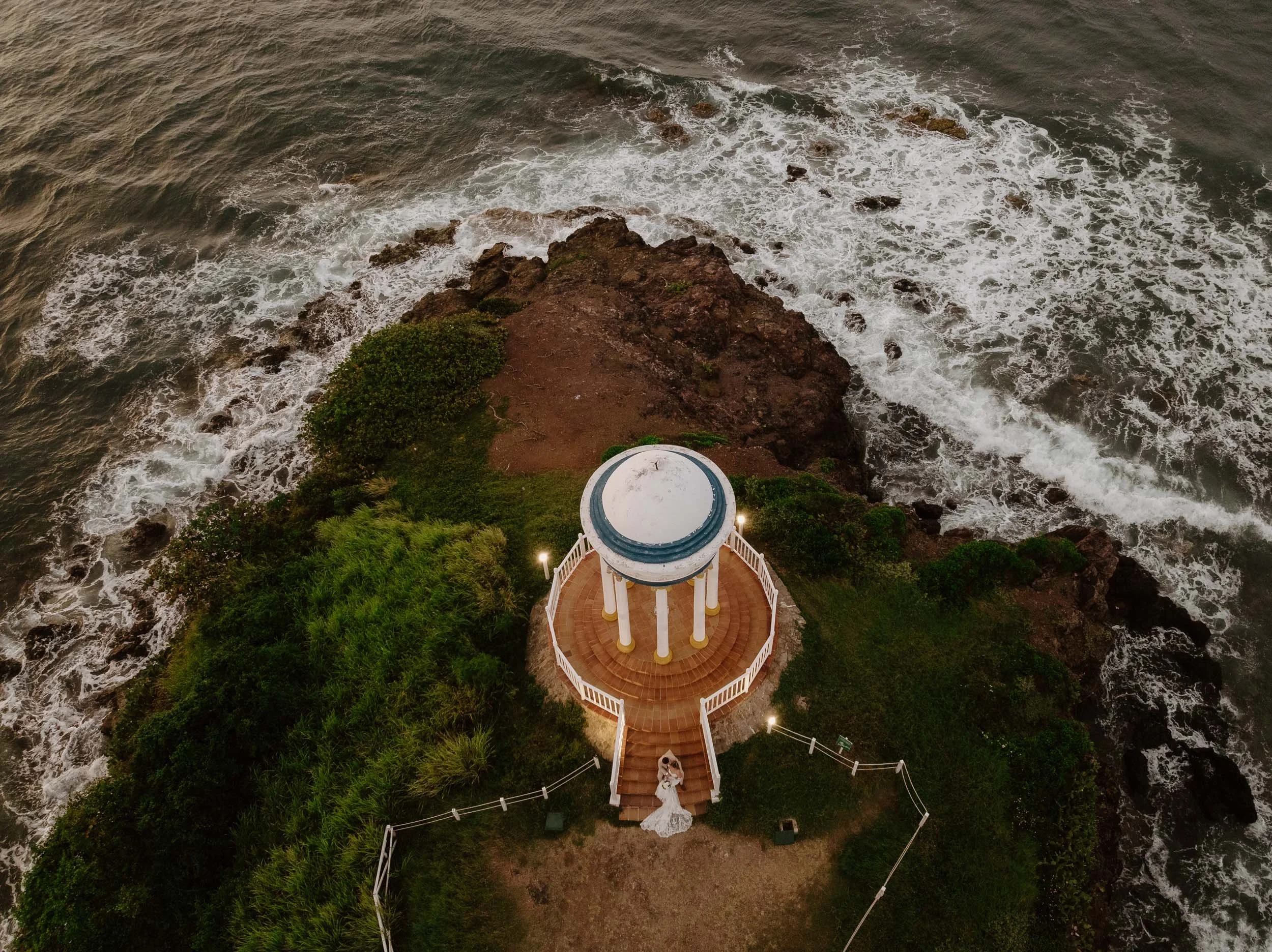 Drone aerial view of the couple siting down in the stairs of a chapel in the top of a cliff close to the sea, you can see the waves of the sea.