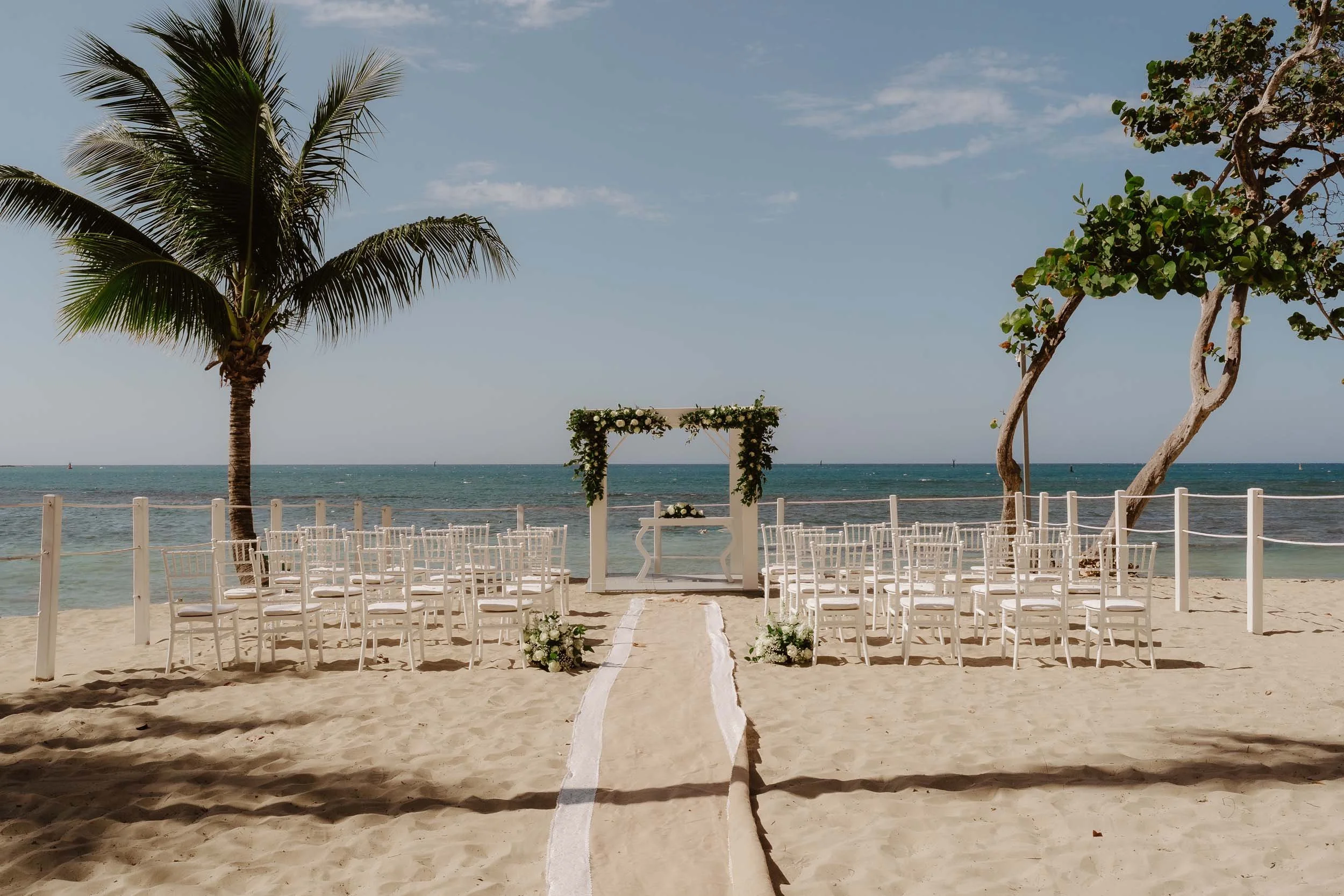 Photo of the ceremony setting. You can see the palm, the greens, the blue sea at the background, the chairs and the wedding aisle.