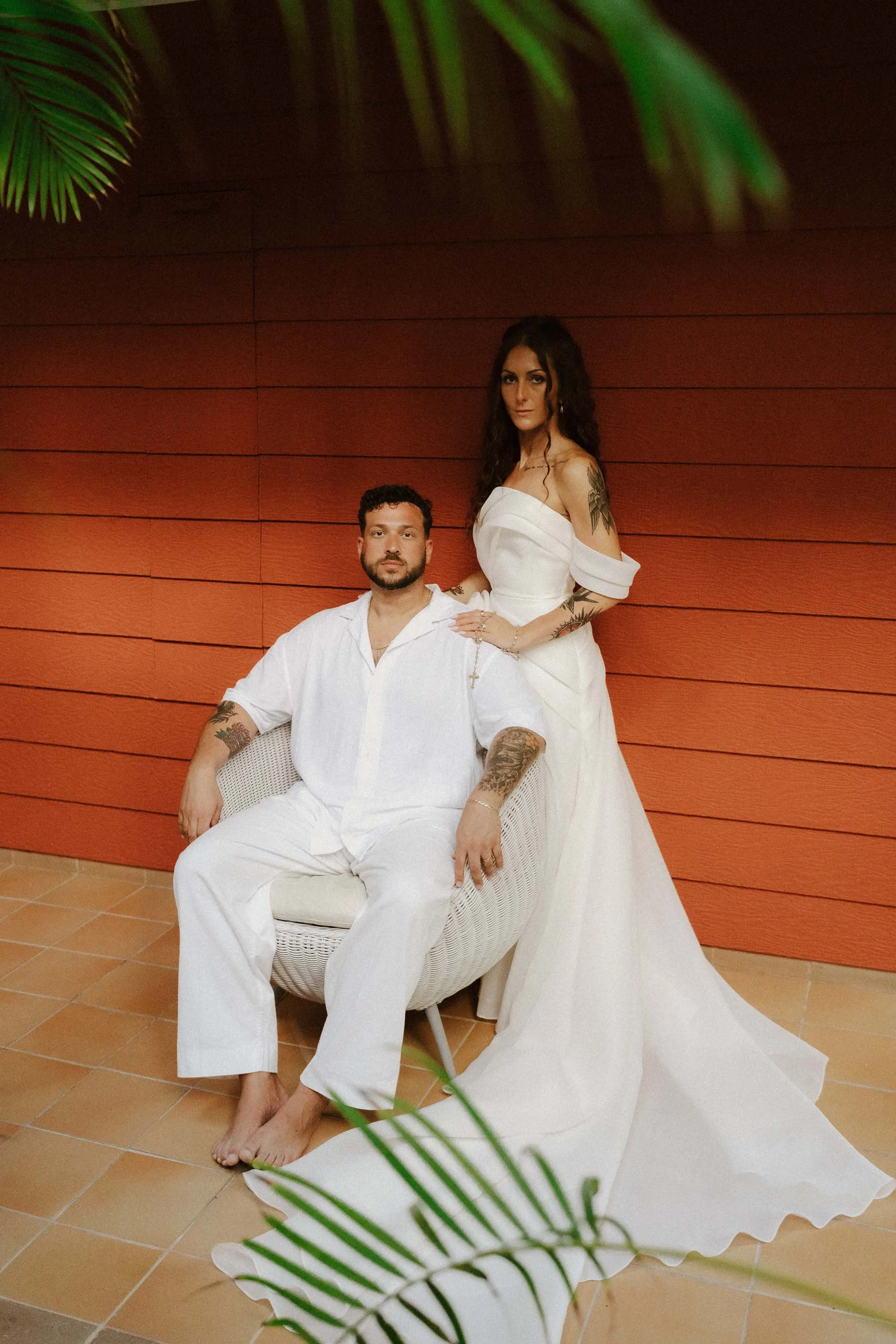 Bride and groom photo. The groom is seating down in a white chair looking to the camera while the bride has her hand on his shoulder by his side. There is a red wooden background from the walls of the resort building