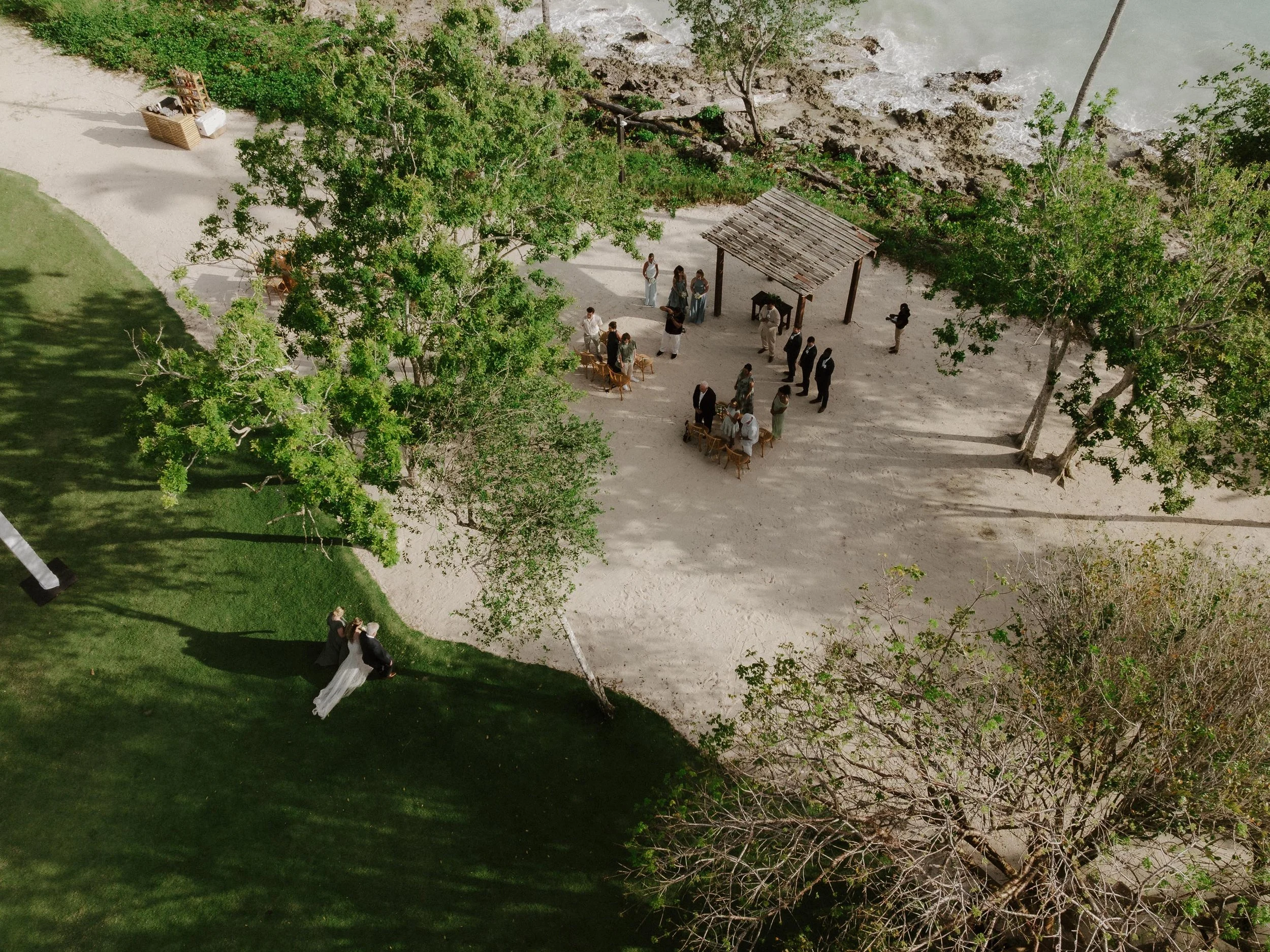 drone photo of the bride entering the ceremony wedding aisle, while the guests and the groom look at her walking down the aisle. You can see the beach, the ocean and the grass
