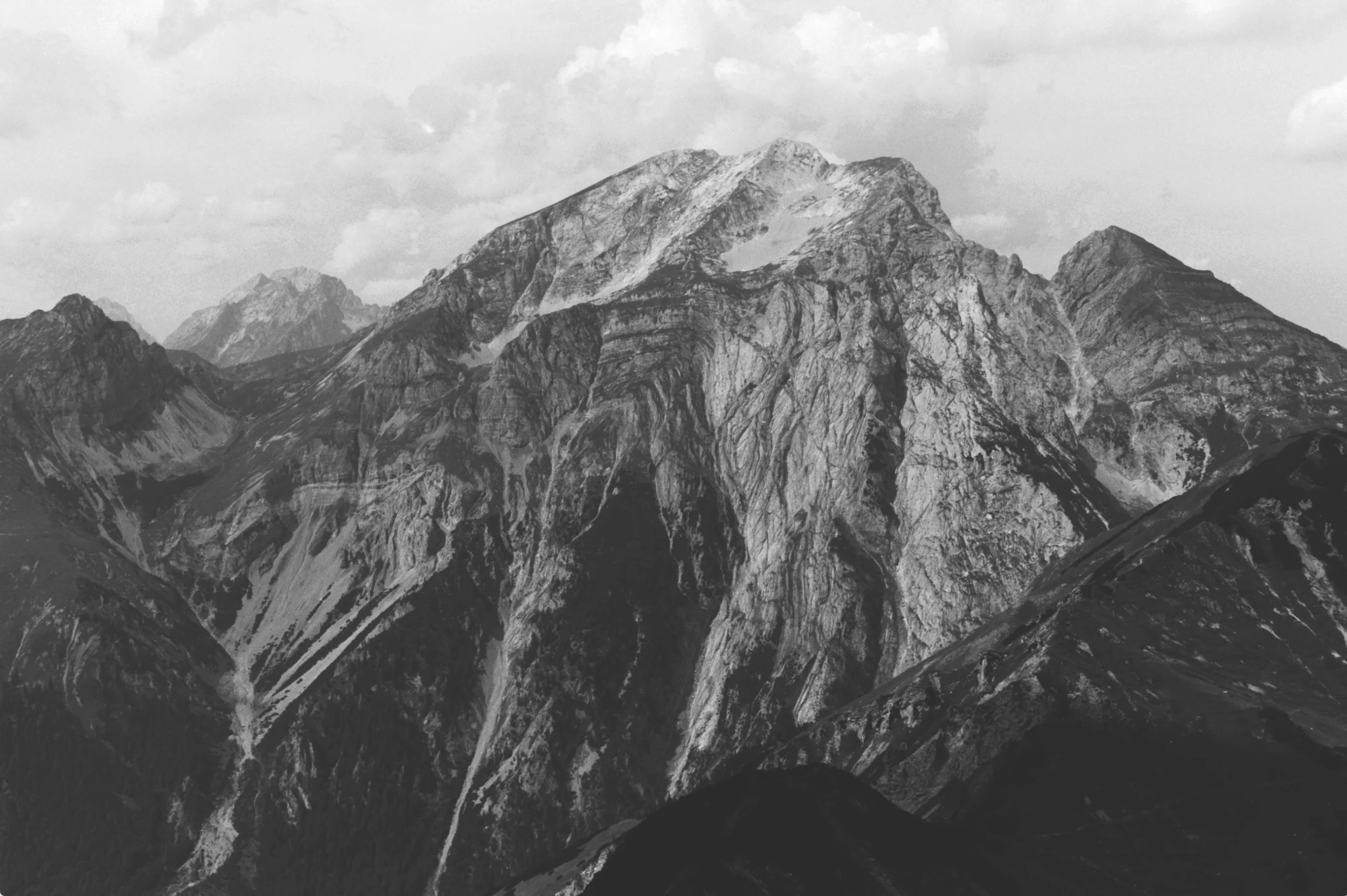 Schwarze und weiße Aufnahme eines Bergmassivs mit hohen Gipfeln und steilen Felswänden unter einem bewölkten Himmel.