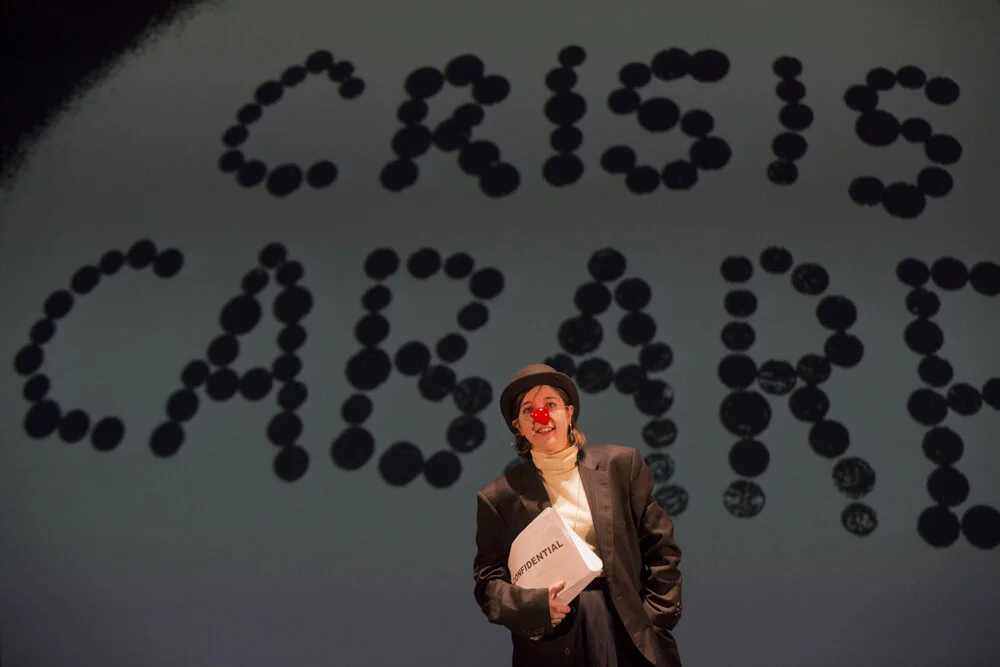Besuited woman with red nose and bowler hat holds papers marked confidential in front of Crisis Cabaret in big letters