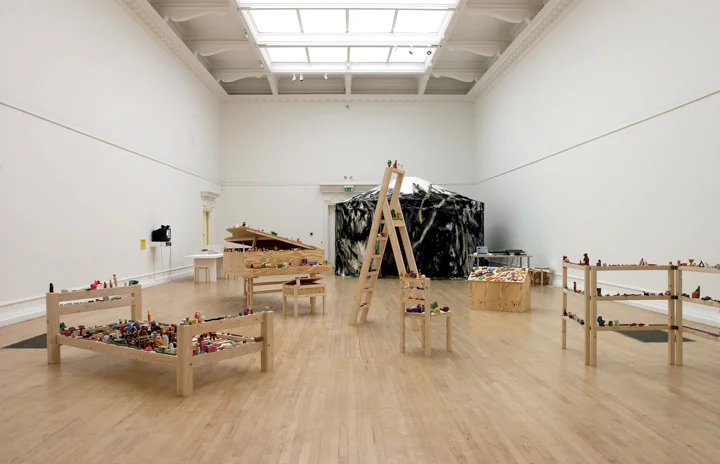 Many small coloured objects on wooden objects - bed, ladder, shelves - in a large art gallery room