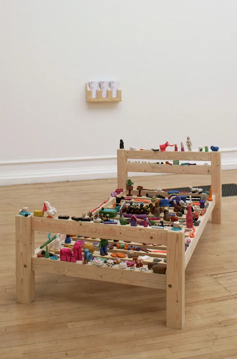A wooden bed frame on which are perched many small brightly coloured plasticine objects
