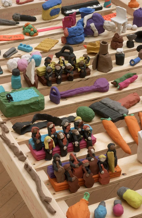 A close-up of a bed frame are perched many plasticine objects including carrots, figures, a handbag