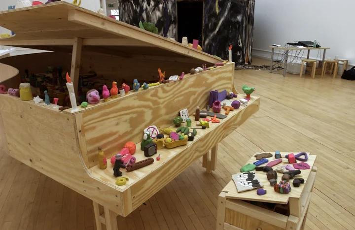 A wooden piano and piano stool on which are perched many small brightly coloured plasticine objects