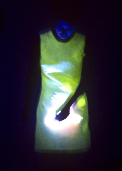 A sleeveless dress worn by Hayley Newman glowing green in darkness, Hayleys face is visible in blue light