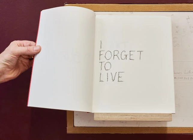 Hand holding open the pages of a book on the right page of which are the words in handwriting - I forget to live