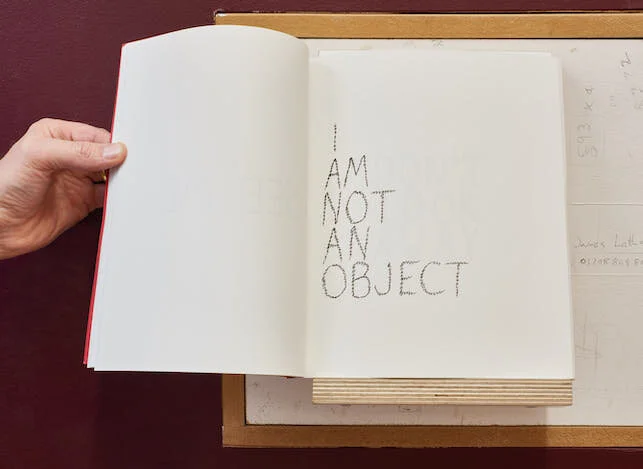 Hand holding open the pages of a book on the right page of which are the words in handwriting - I am not an object