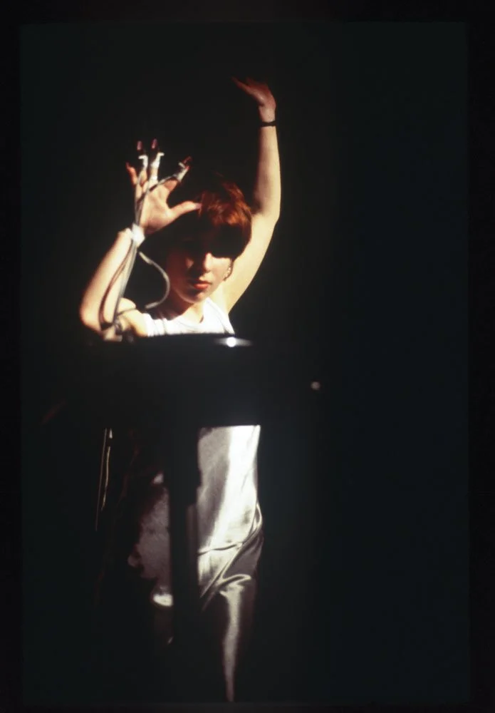 Hayley Newman in a silver sleeveless dress gestures upwards with wires attached to her right hand