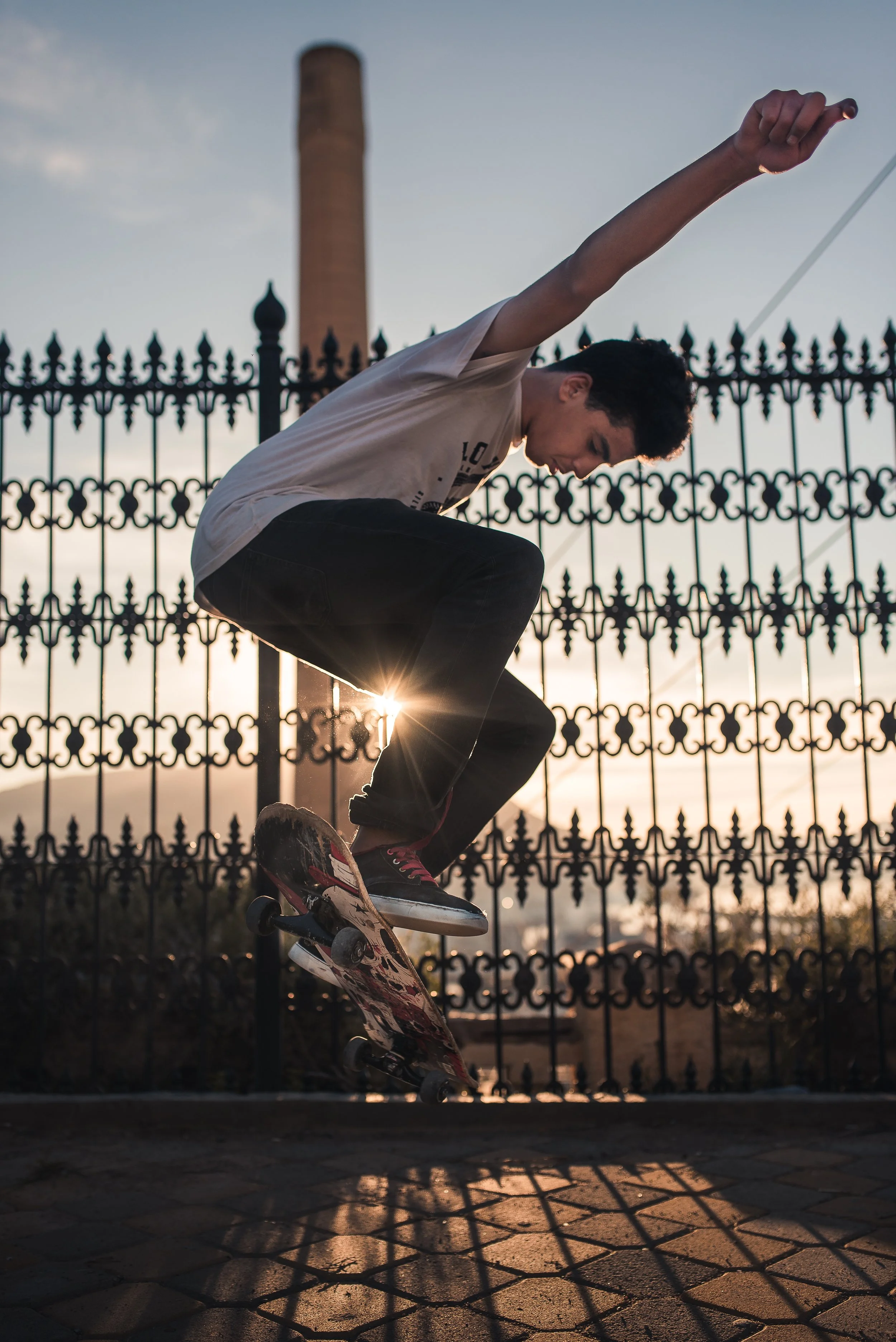 A young man performing a skateboarding trick at sunset, crouched with arms extended, in front of a decorative iron fence and a tall brick column, with long shadows cast on the paved ground.