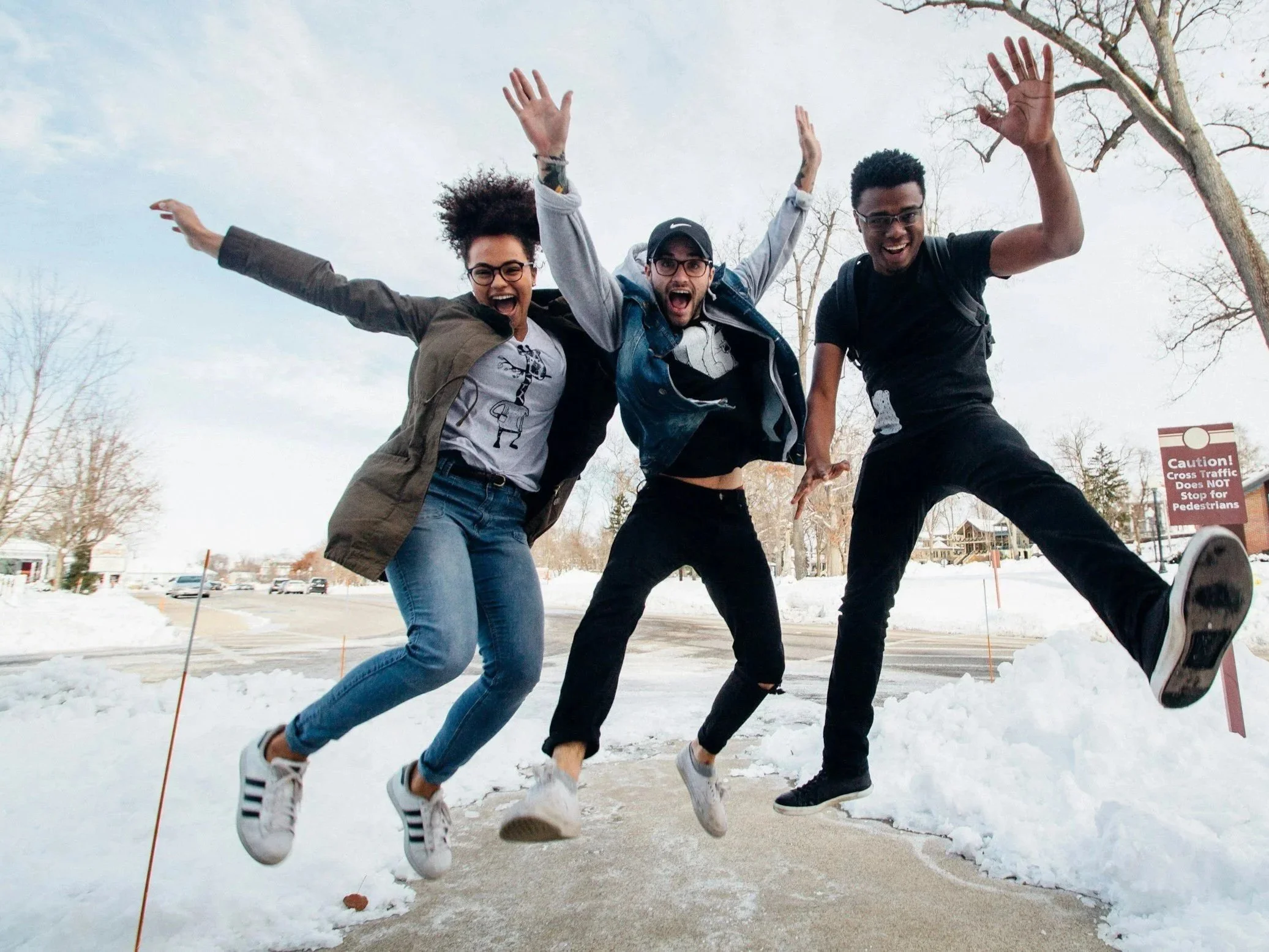 Three young adults jumping in the snow in a wintery outdoor setting. They are smiling and appear to be having fun. There is snow on the ground and leafless trees in the background.