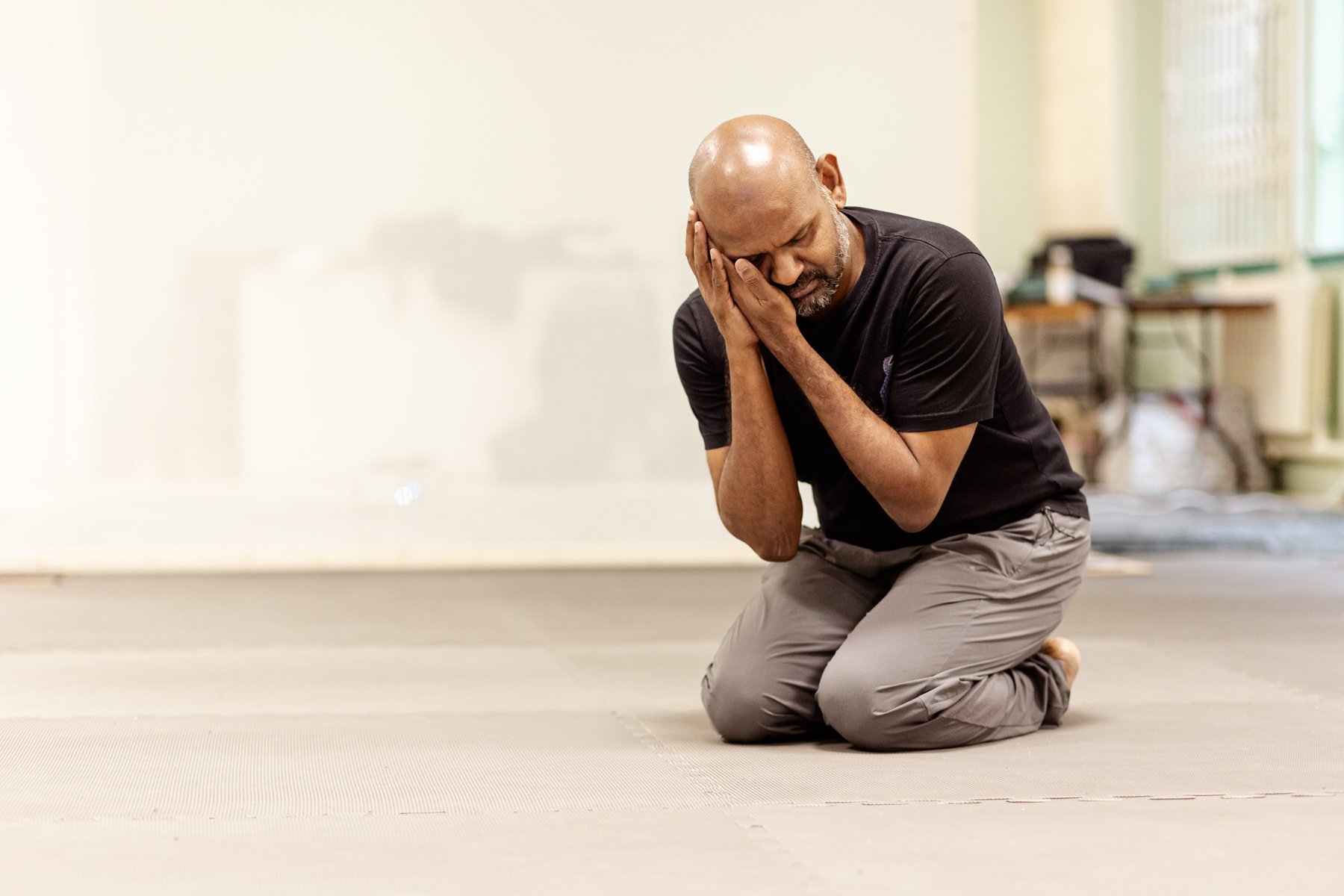  Photo of actor Ramesh Meyyappan in the rehearsal room. He is kneeling with both of his hands cradling his face. His eyes are closed and his head is slightly turned and resting on his hands. 