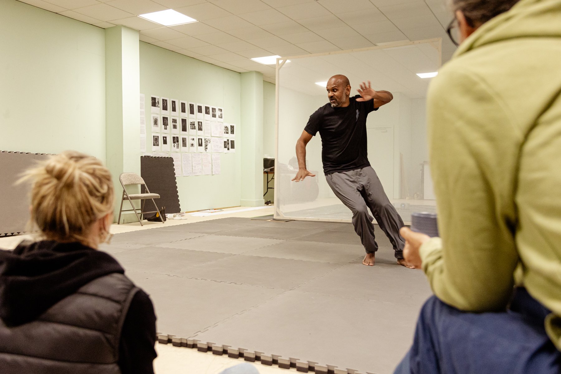  Photo of actor Ramesh Meyyappan in the rehearsal room in the background, in the middle of a dynamic movement across the floor. In the foreground are the backs of two people watching him. Lots of pages of images and writing are stuck on the wall. 