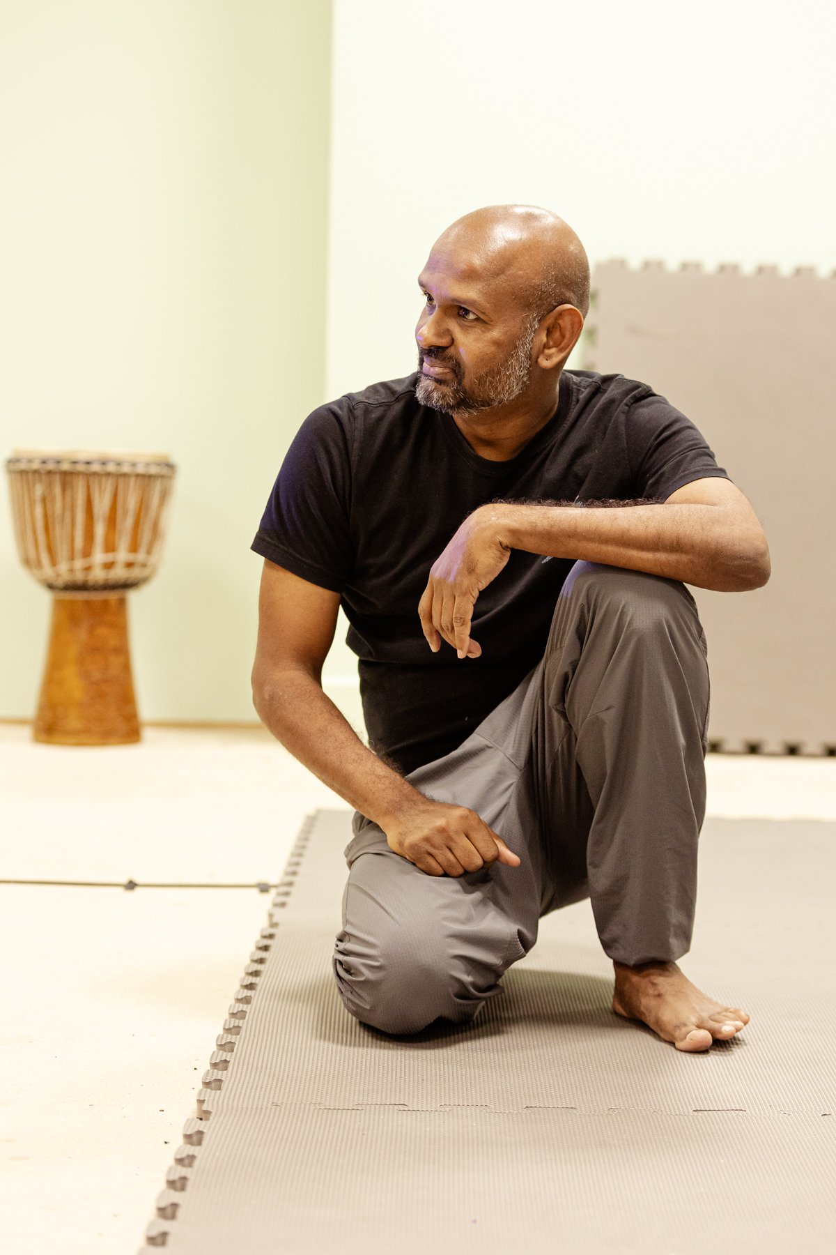  Photo of actor Ramesh Meyyappan in the rehearsal room. He is kneeling on one knee in a relaxed position and is smiling at something off camera. 