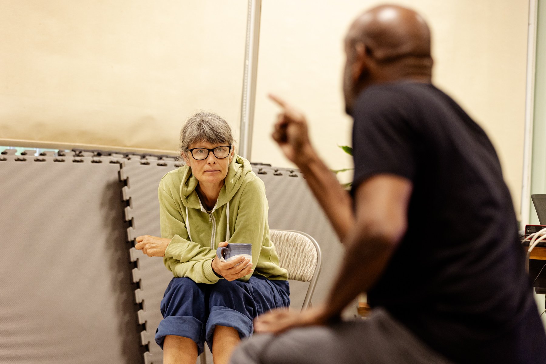  Photo of designer Katie Sykes sitting looking with concentration at actor Ramesh Meyyappan in the rehearsal room, who is slightly blurry in the foreground and pointing his finger. 