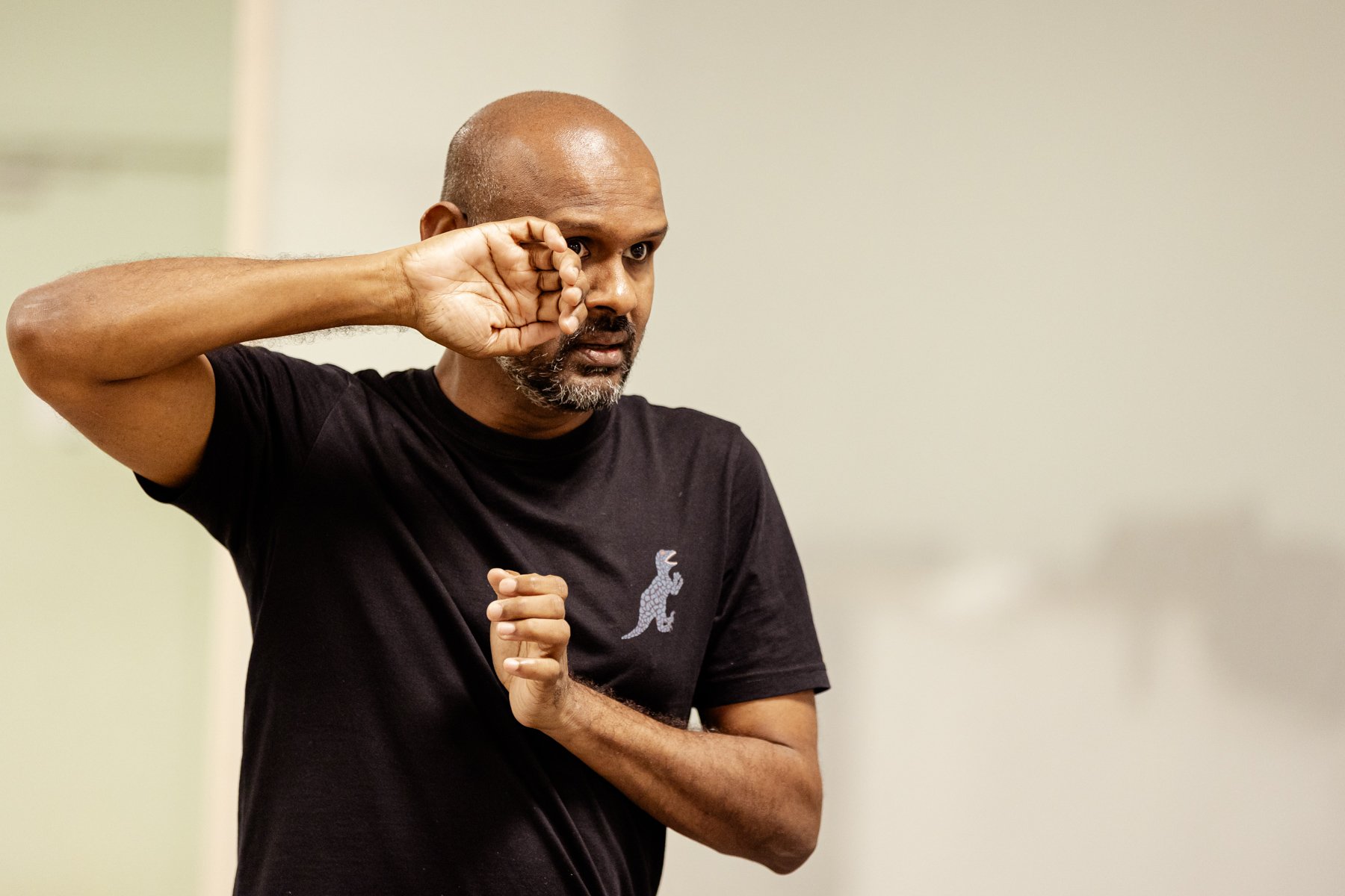  Photo of actor Ramesh Meyyappan in the rehearsal room. He has his hands raised and is miming pushing something to one side to look around it. 