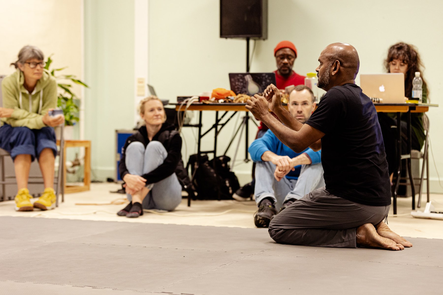  Photo of people in the rehearsal room for Last Rites. Three people are sitting on the floor and two people are sitting behind a table with laptops. Everyone is looking at actor Ramesh Meyyappan who is kneeling and in the middle of acting something o