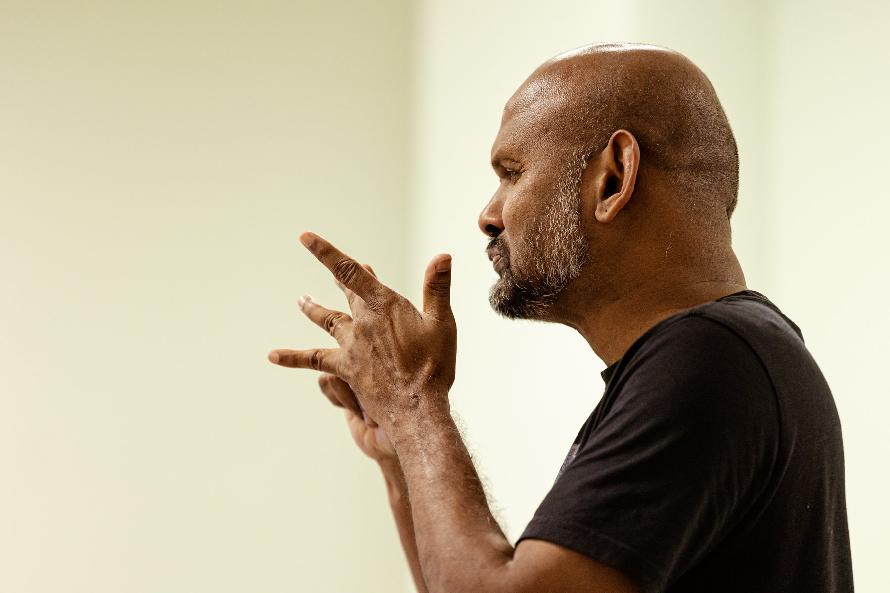 Photo of actor Ramesh Meyyappan in the rehearsal room. He is facing sideways and is in the middle of signing something with his hands, his fingers are raised. 