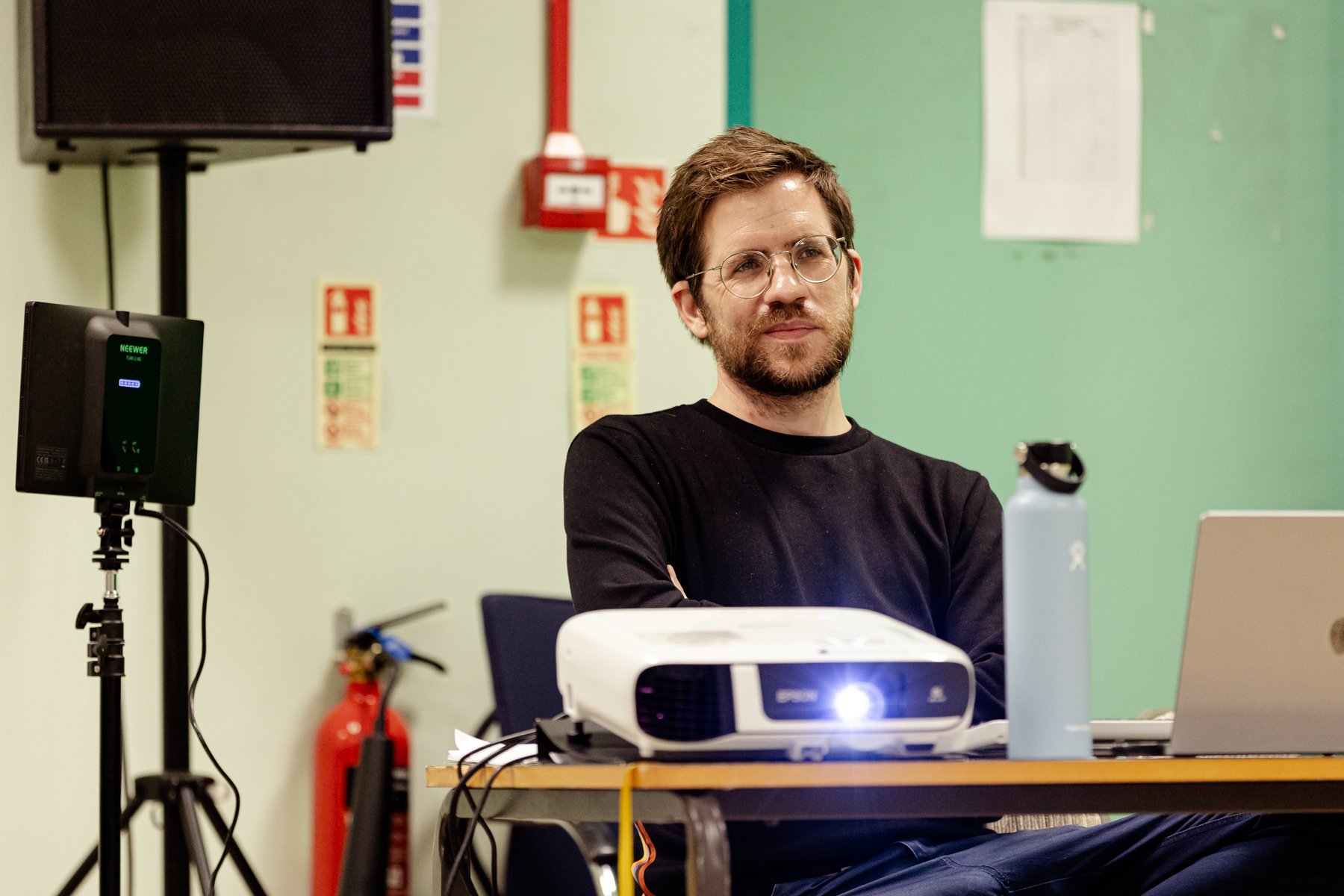  Photo of video and projection designer Chris Harrisson sat at a table with a projector in front of him. He is looking at something off camera and smiling. 