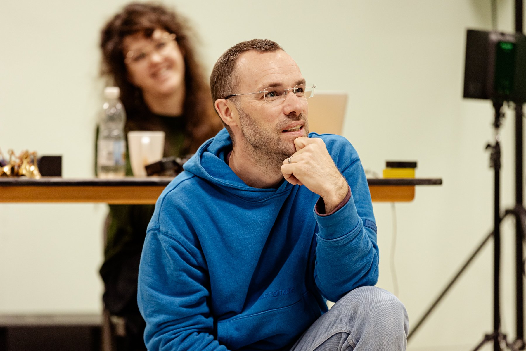  Photo of director George Mann and stage manager Clare Hannah in the rehearsal room. George is in the foreground and wearing a blue hoodie. He is kneeling on the floor with one hand under his chin and smiling up at something off camera. Hannah is sli