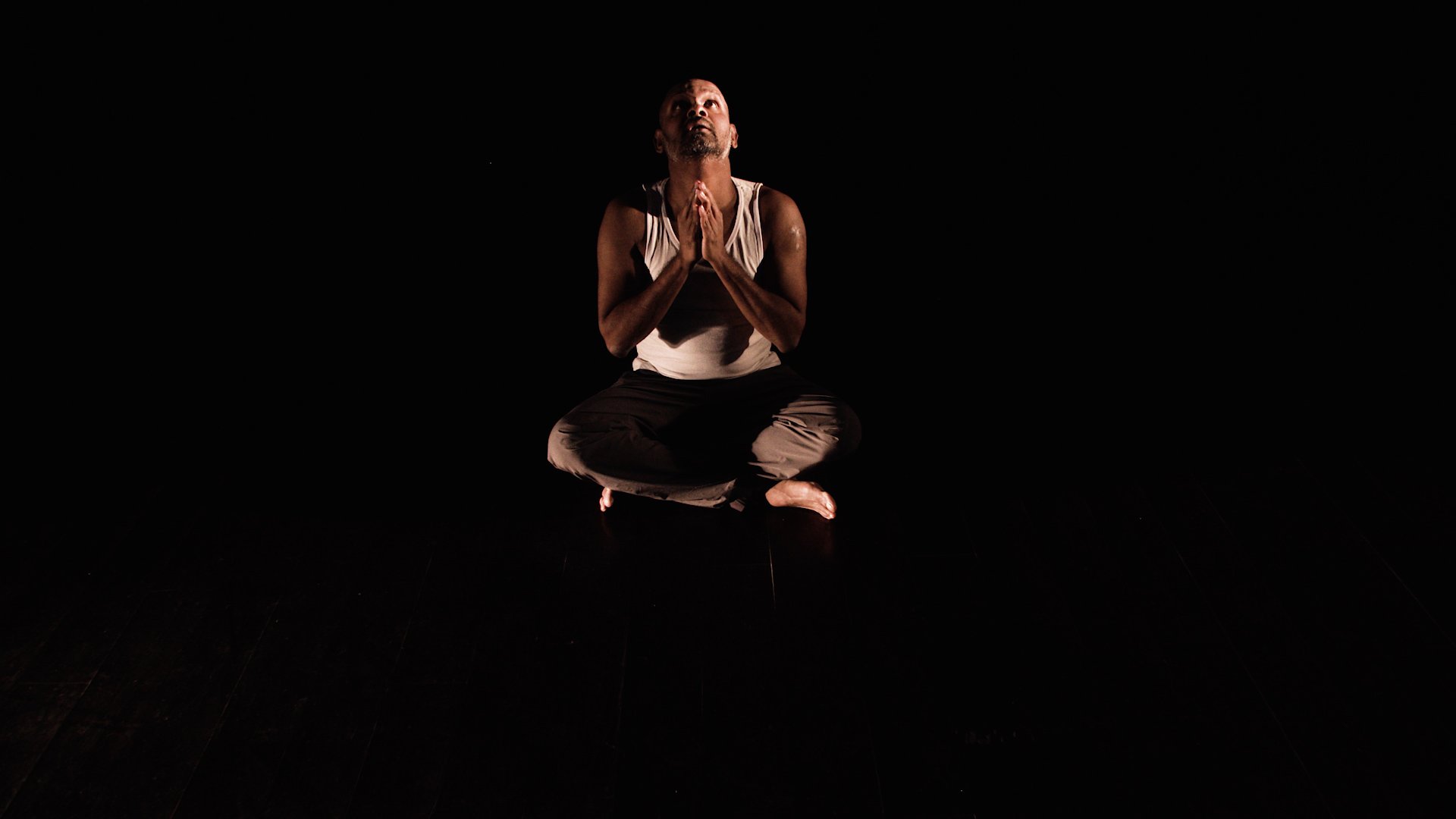  Photo of actor Ramesh Meyyappan. He is sat cross legged with his hands in a prayer position, looking up. He is sitting on a black floor with a black background so it looks as though he is floating! 