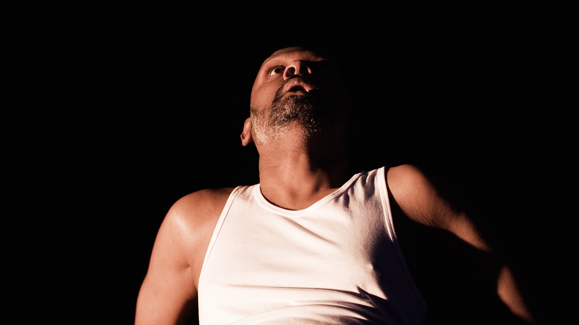  Photo of actor Ramesh Meyyappan. He is wearing a white vest and the image shows his chest and head. He is against a black background and looks like he is falling backwards. He is looking up above him with his mouth slightly open. 