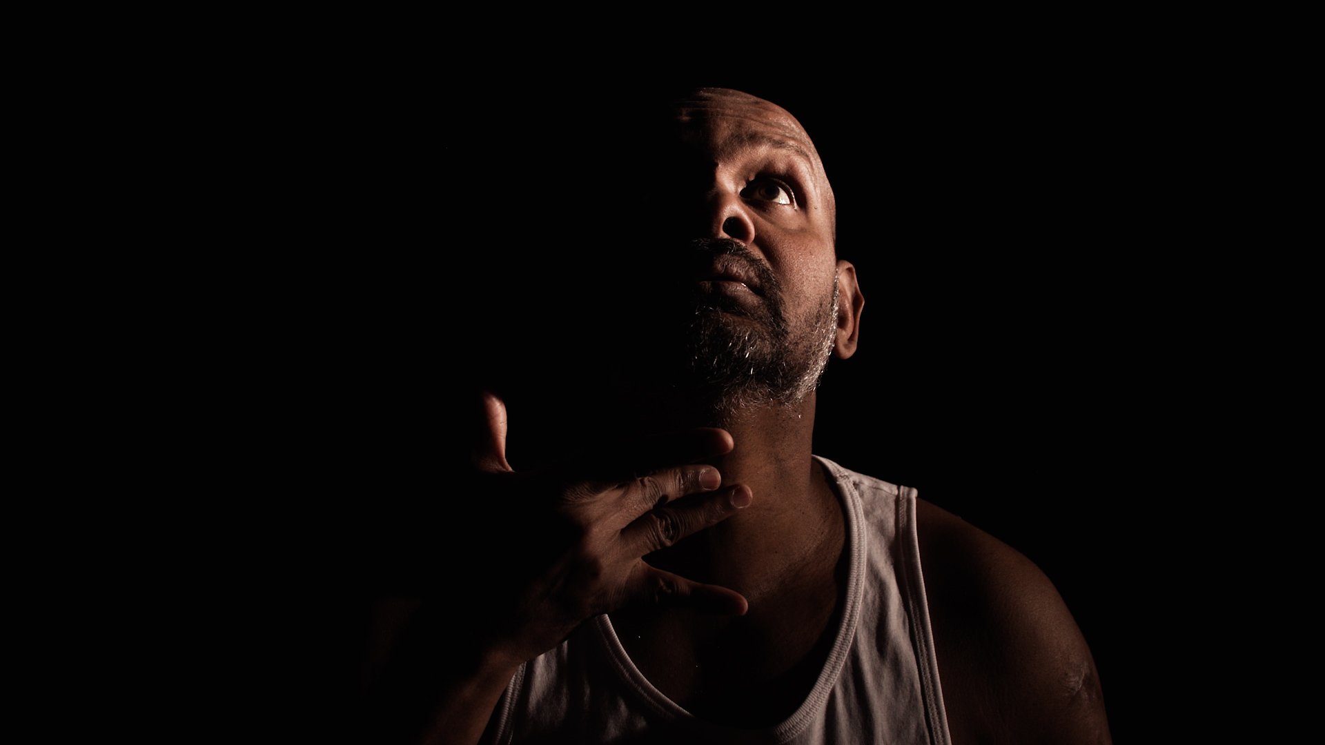  Photo of actor Ramesh Meyyappan against a black background. He is lit from the side so the right hand side of his face and shoulders are in shadow. He is looking upwards and his fingers are touching his throat. 
