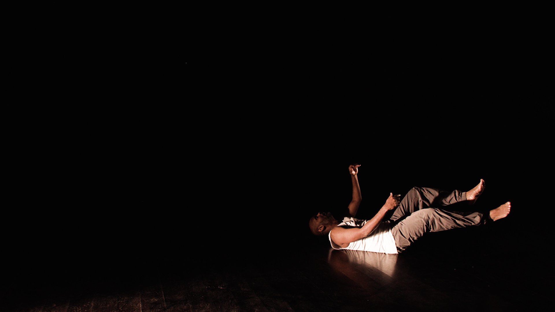  Photo of actor Ramesh Meyyappan. He is to the right hand side of the image and wearing a white vest and grey trousers. He is against a black background and sparsely lit. He appears to have just fallen onto his back and has his arms and legs raised. 