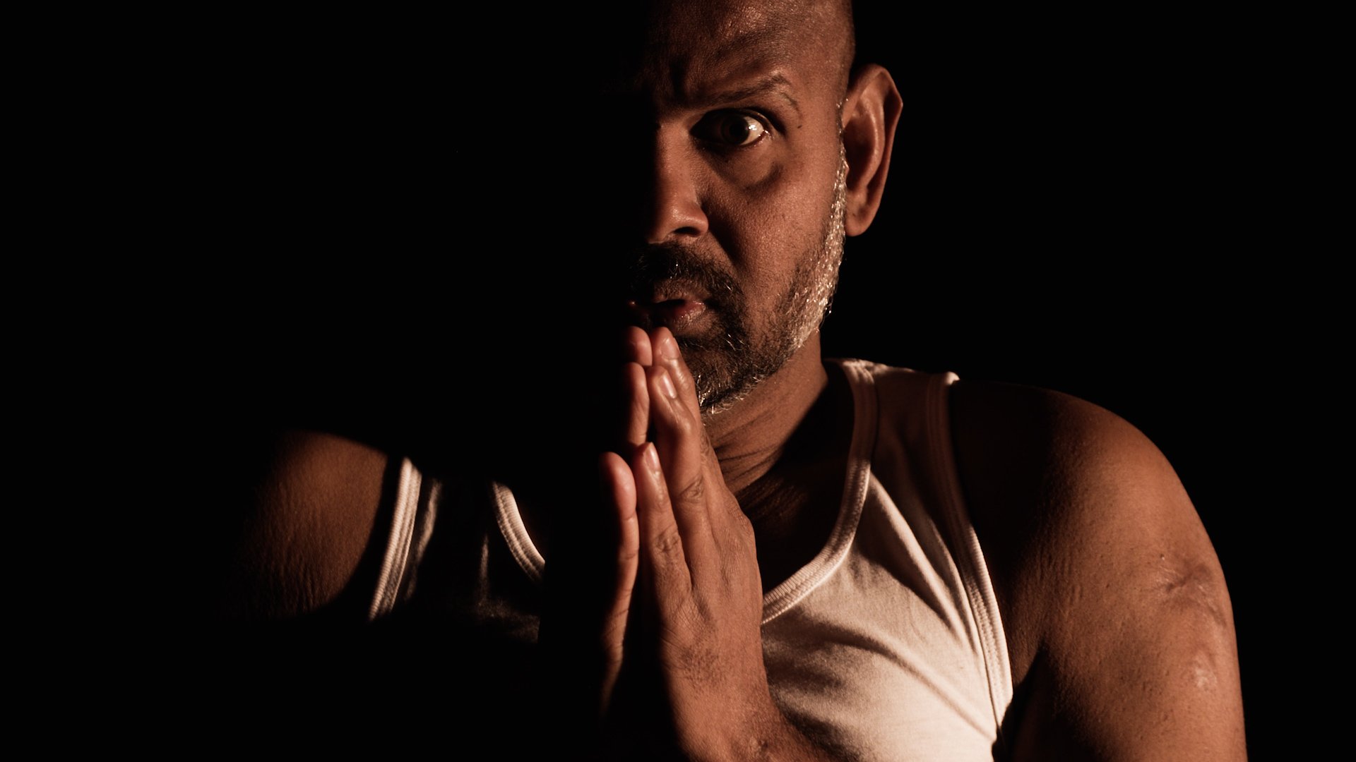  Photo of actor Ramesh Meyyappan wearing a white vest. Only his head and shoulders are visible and he is lit from the side so his right hand side is in shadow. His eyes are fairly wide open and his hands are raised in a prayer position. 