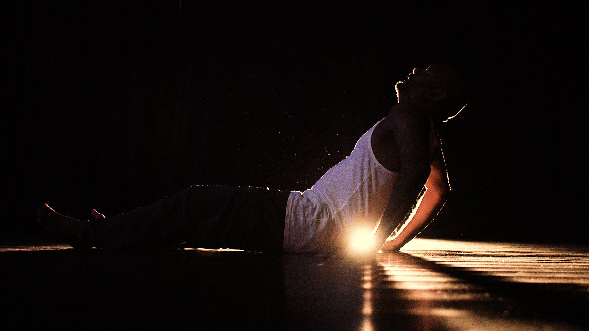  Photo of actor Ramesh Meyyappan. The image is mostly dark, with a single bright light shining towards the camera at the centre. This is back-lighting Ramesh who is sat on the floor holding himself up on his hands and looking upwards. 