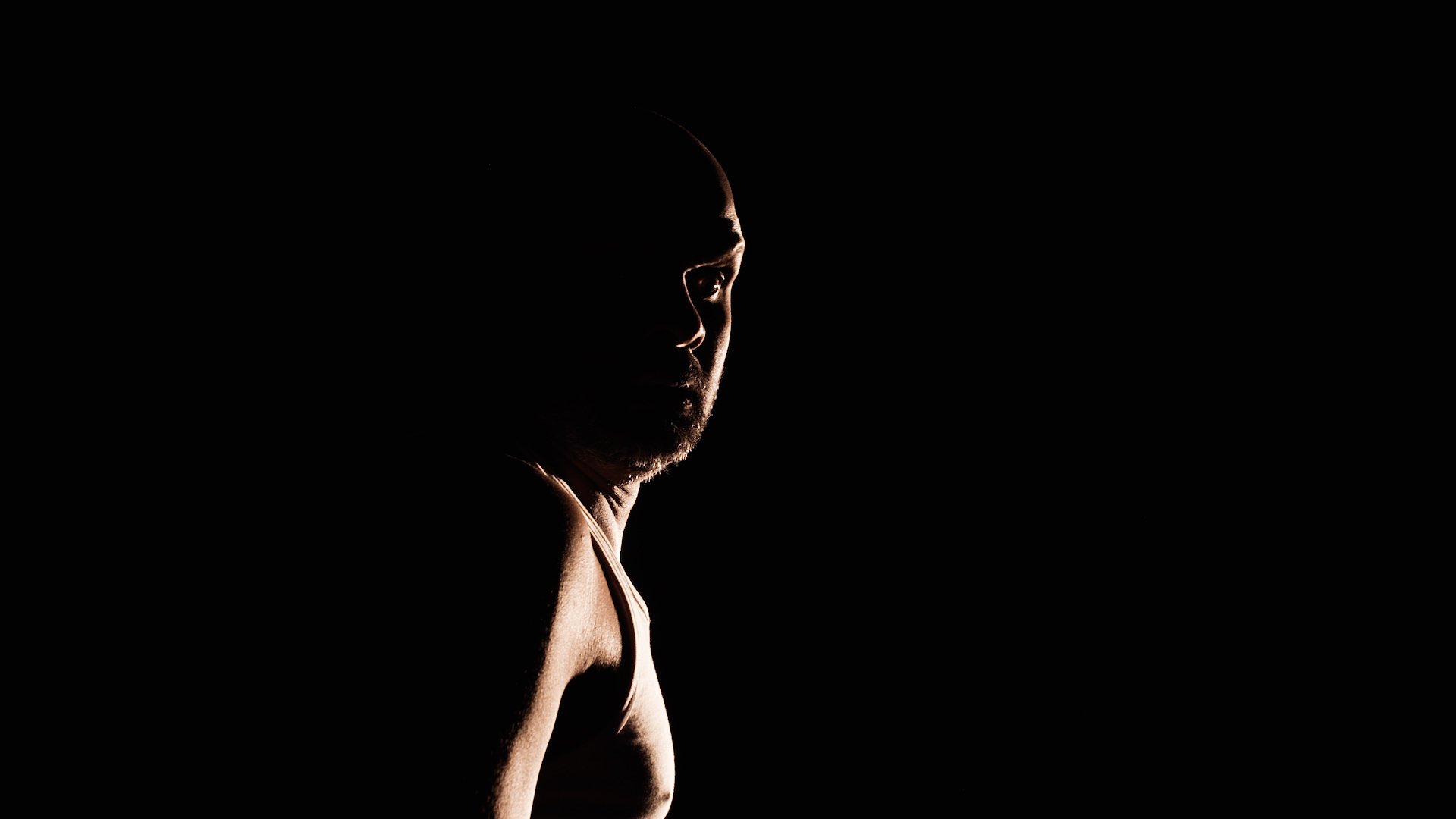  Photo of actor Ramesh Meyyappan standing sideways looking towards the camera over his right shoulder. He is mostly in shadow and you can just make out the profile of his face and chest. 