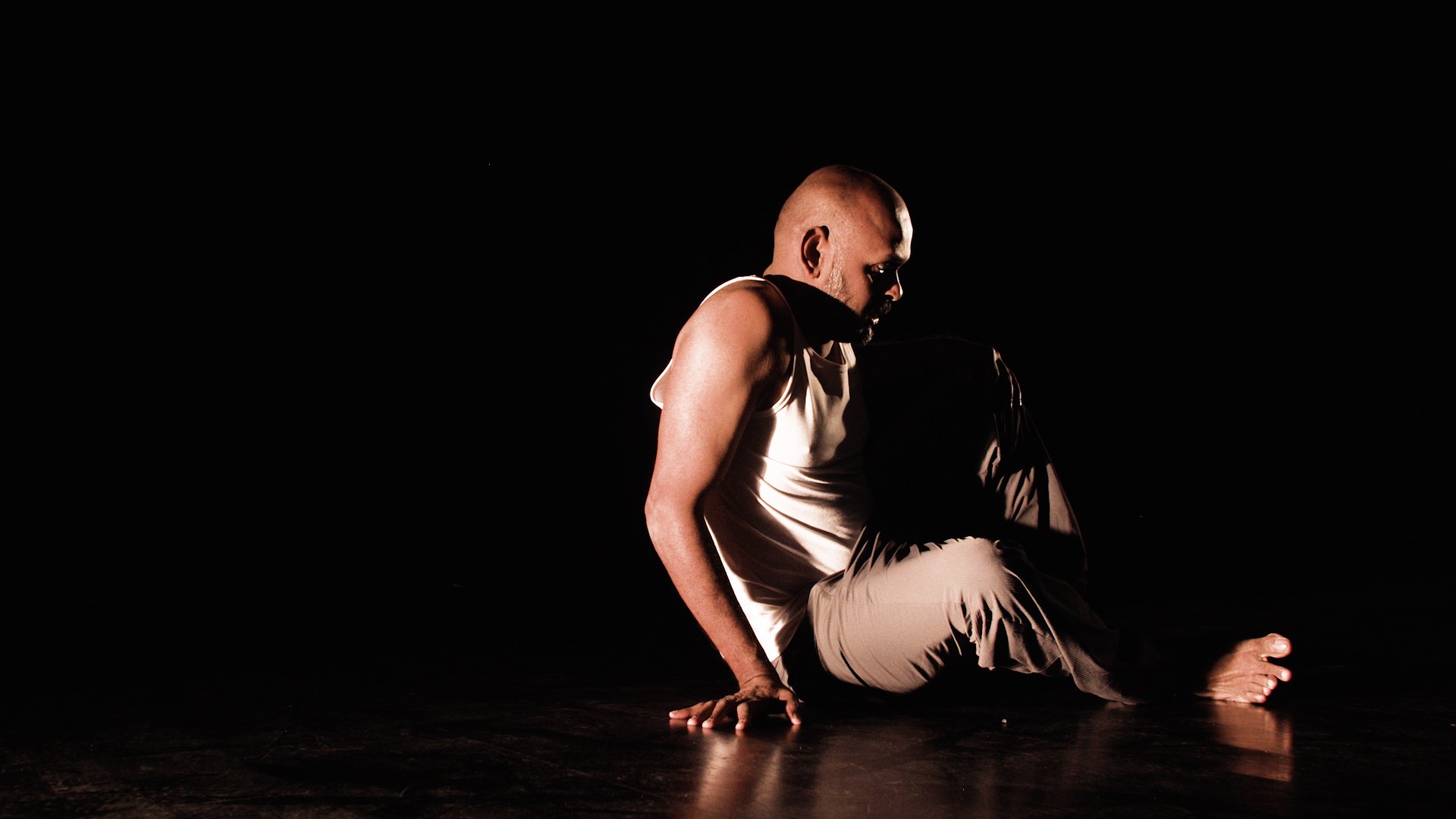  Photo of actor Ramesh Meyyappan sitting on the floor looking as though he is in the middle of getting up. The background and floor is black and Ramesh is wearing a white vest and grey trousers. He is partly in shadow. 