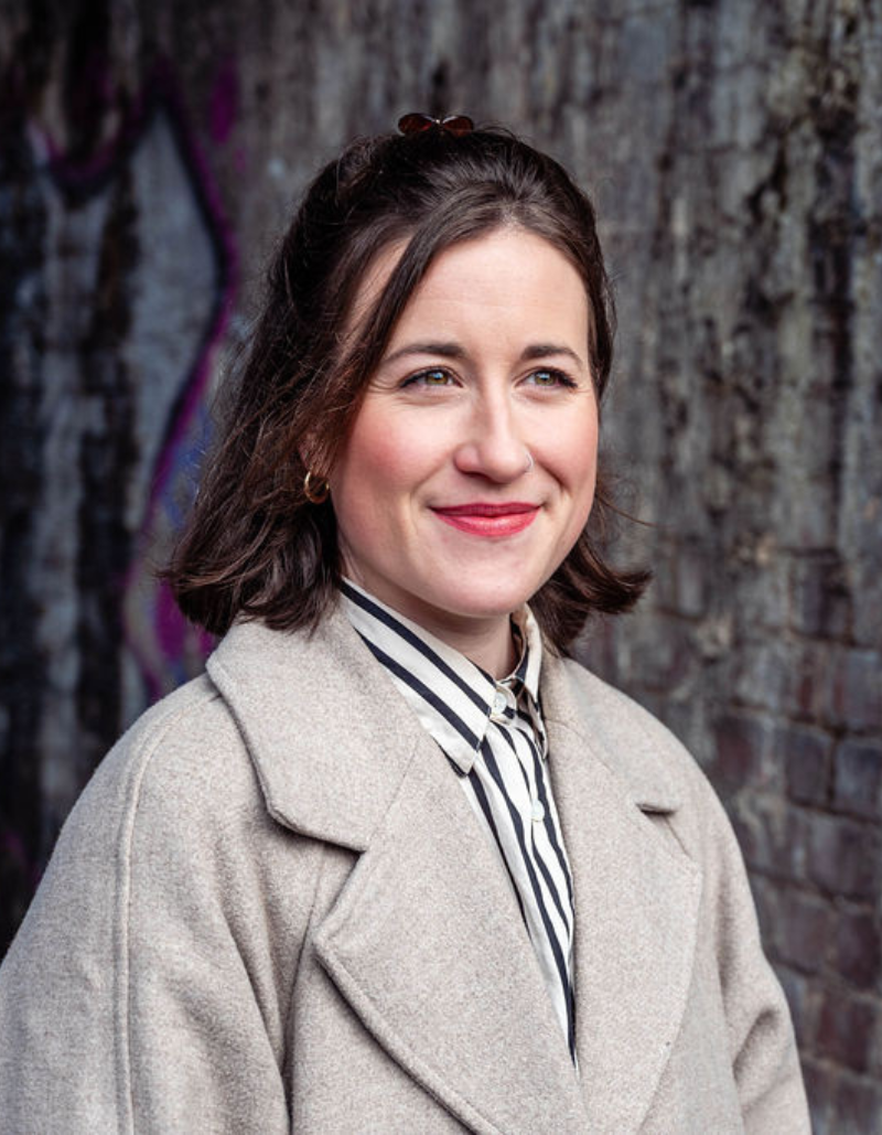 Photo of Ali Ford who has short haird clipped back, is wearing a stripey blouse and a grey coat and is smiling looking to the right of the camera.