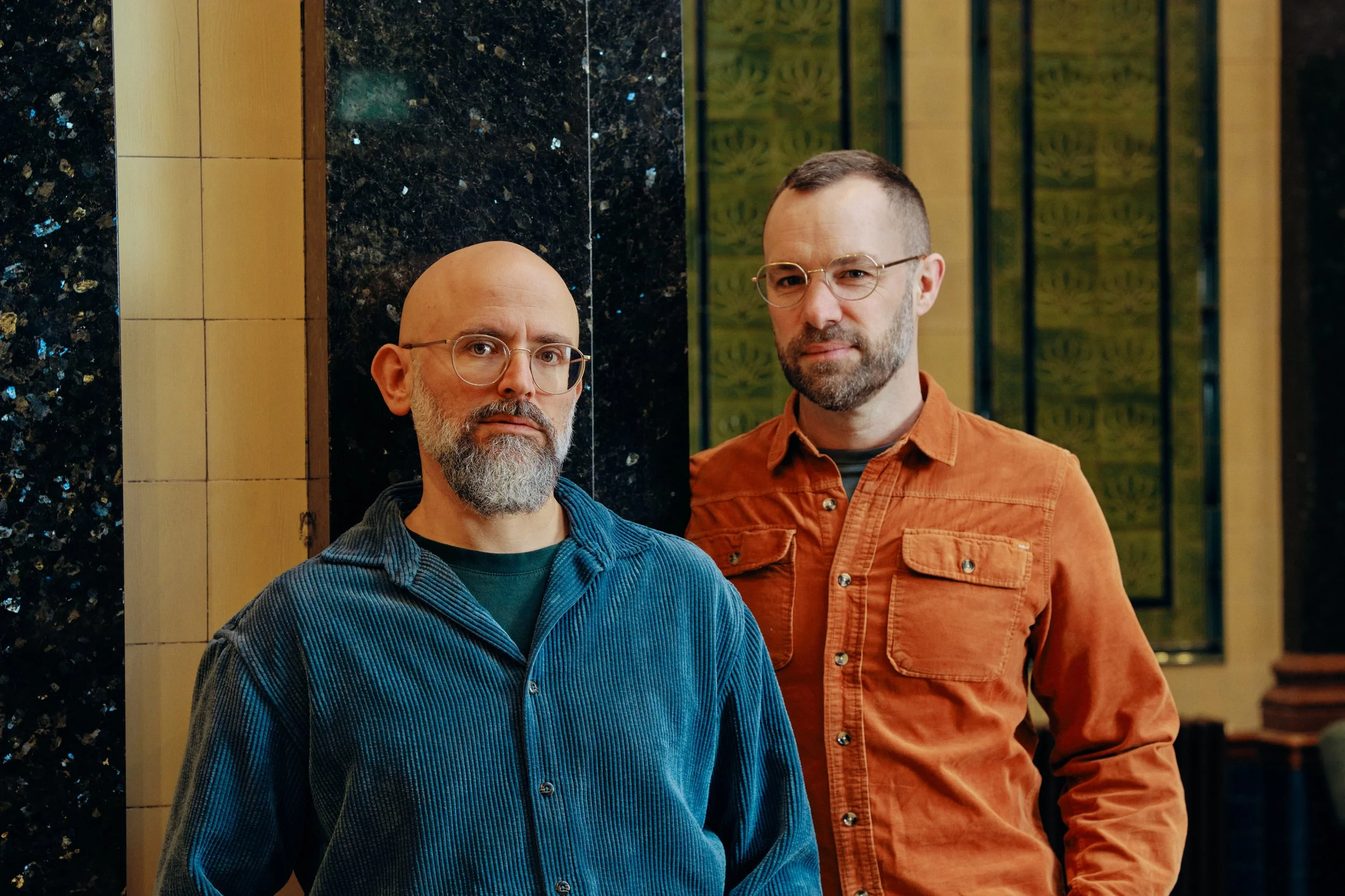 Photo of Co-Artistic Directors Nir Paldi (stood on the left with a beard and glasses, wearing a turquoise shirt) and George Mann (stood on the right with a beard and glasses, wearing an orange shirt). Both are looking straight at the camera.