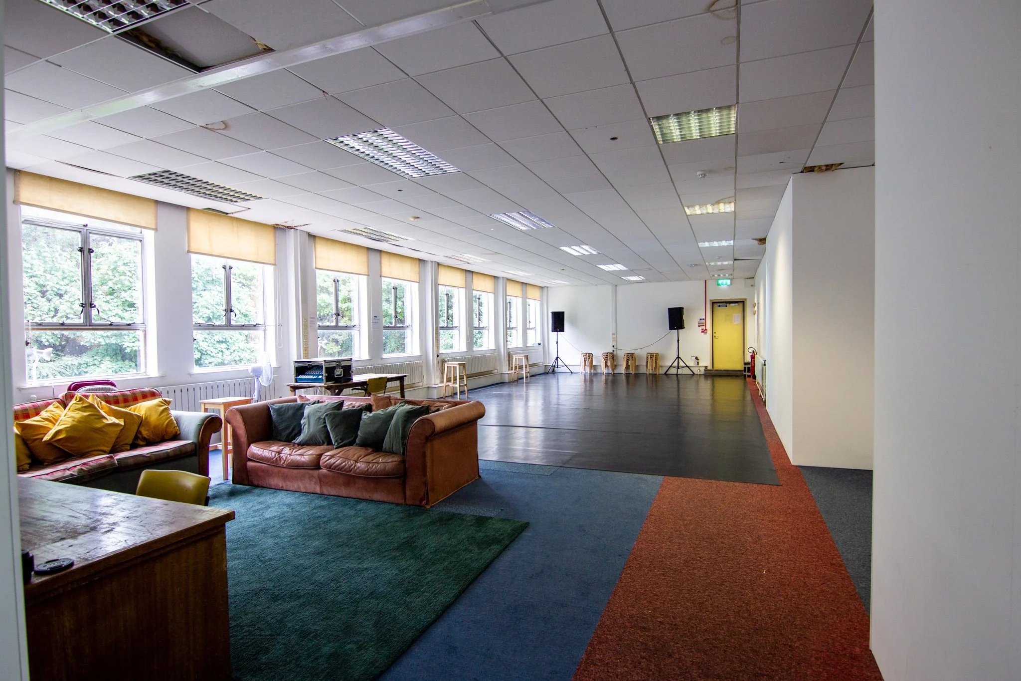 A large room with white walls with seven windows on the left. A sofa in the foreground and a stack of stools in the background. In the foreground there is a rug, whereas the rest of the space is mostly black dance floor.