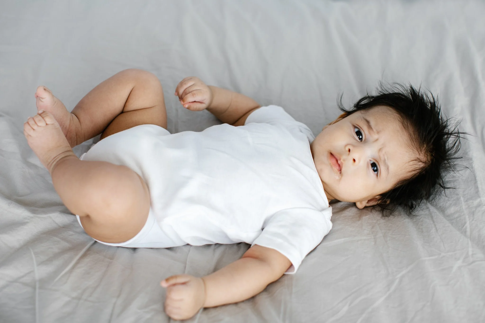 En bebis ligger på rygg på en ljusgrå säng, klädd i en vit body, med mörkt, ostyrigt hår och ser mot kameran.