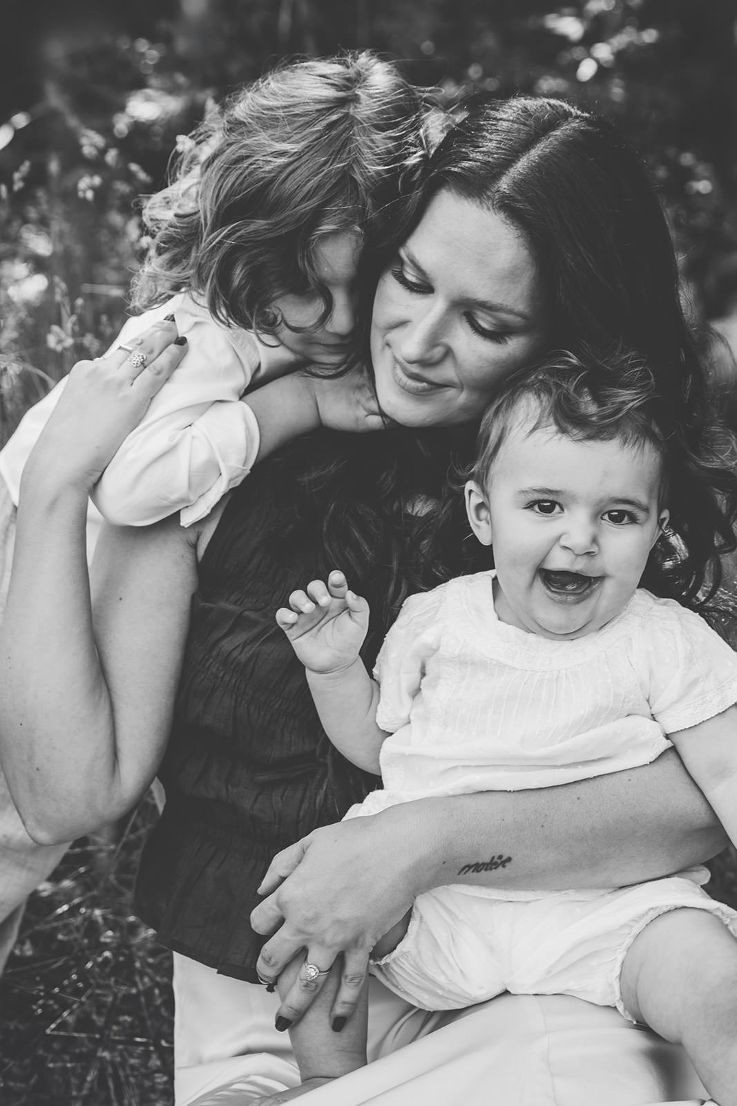 mamma sitter i gräset med sina två barn. Sonen står bakom och håller om henne och dottern sitter i knät.