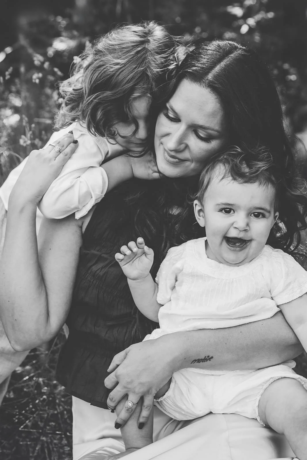 mamma sitter i gräset med sina två barn. Sonen står bakom och håller om henne och dottern sitter i knät.