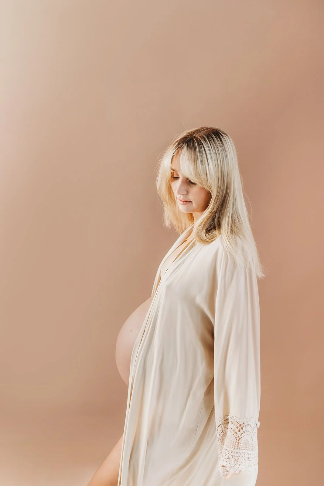 Gravid tjej som står mot beige vägg och fotas. Hon har en beige kimono och visar magen.