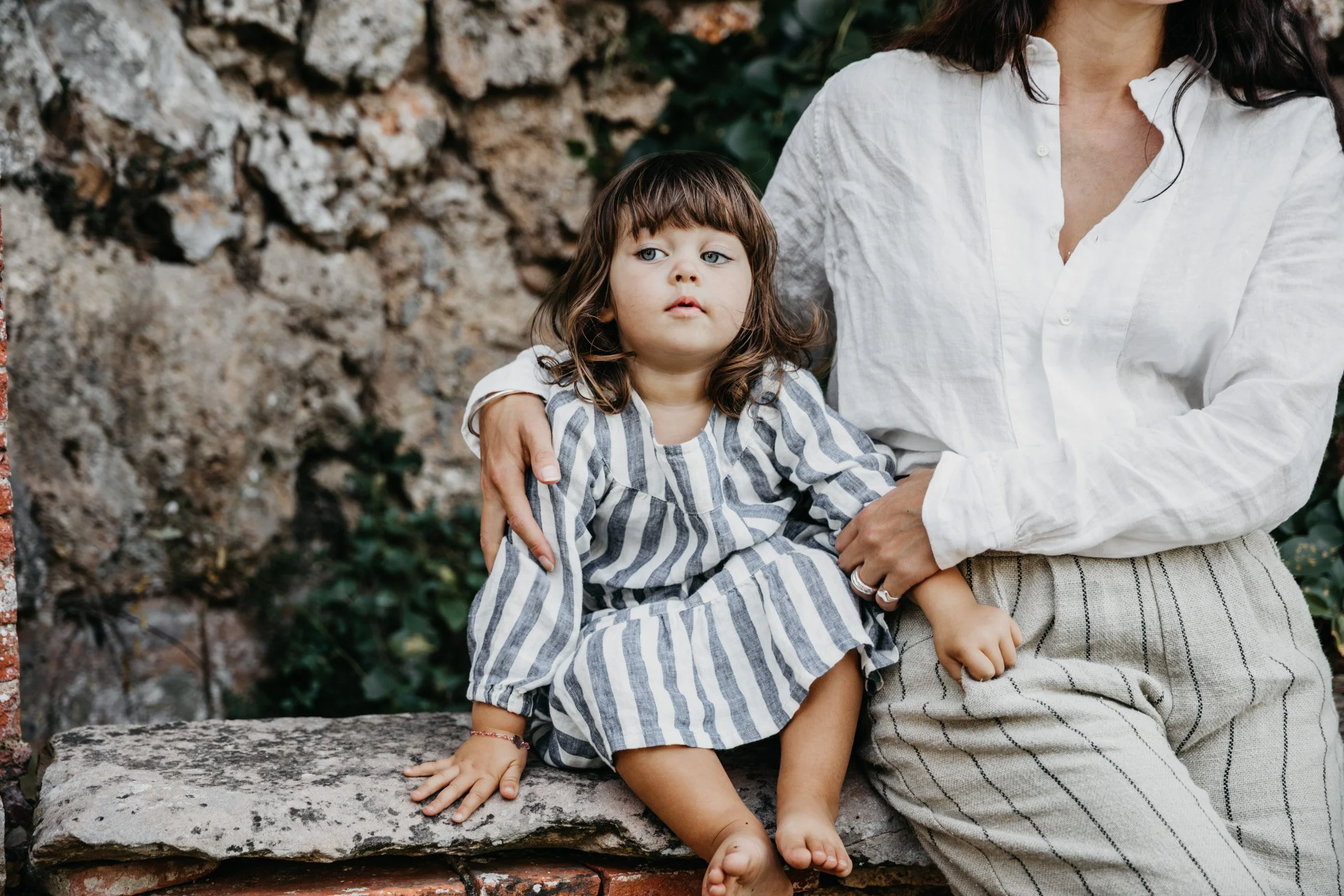 Liten flicka som sitter på en stenmur. Hon har mörkt axellångt hår och en randig klänning