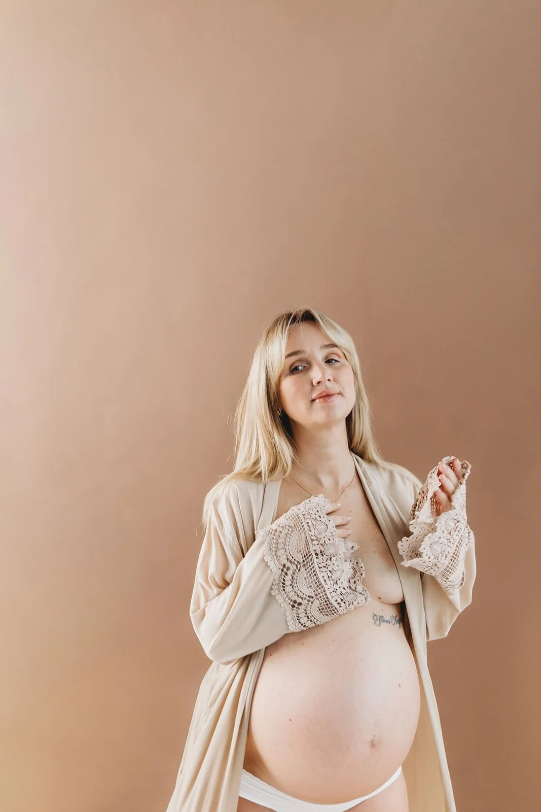 Tjej som står mot brun vägg och fotas och visar upp sin gravidmage. Hon är blond.