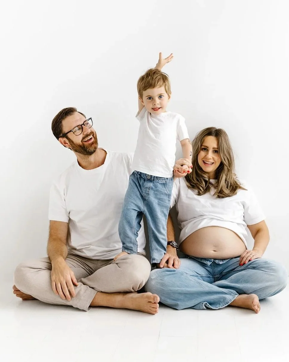 Mamma, pappa sitter på golvet och deras son står i knät på föräldrarna. De har jeans och vita t-shirts.