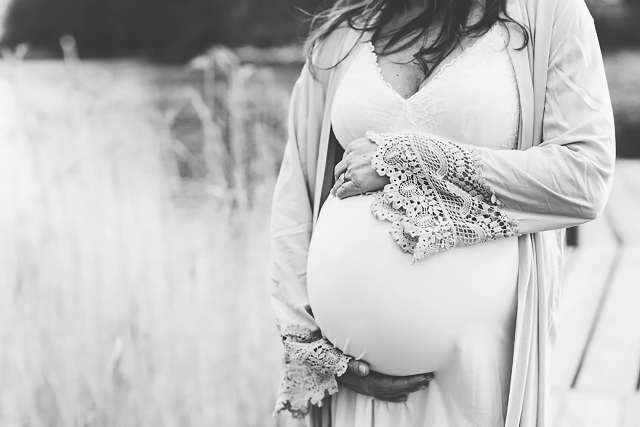 En gravid kvinna i vit klänning med spetsdetaljer, som håller sin mage med båda händer, utomhus.